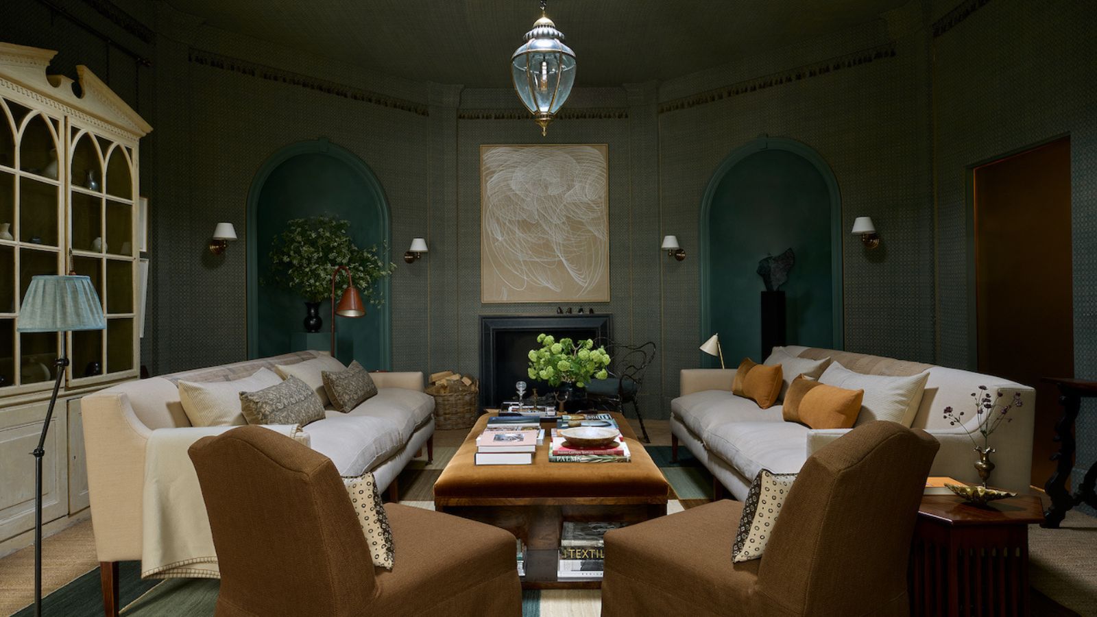 Salon avec murs en toile bleue, alcôves avec statues, table basse en velours marron avec livres dessus, canapés en lin crème, fauteuils marron avec housse, armoire avec livres et suspension en verre