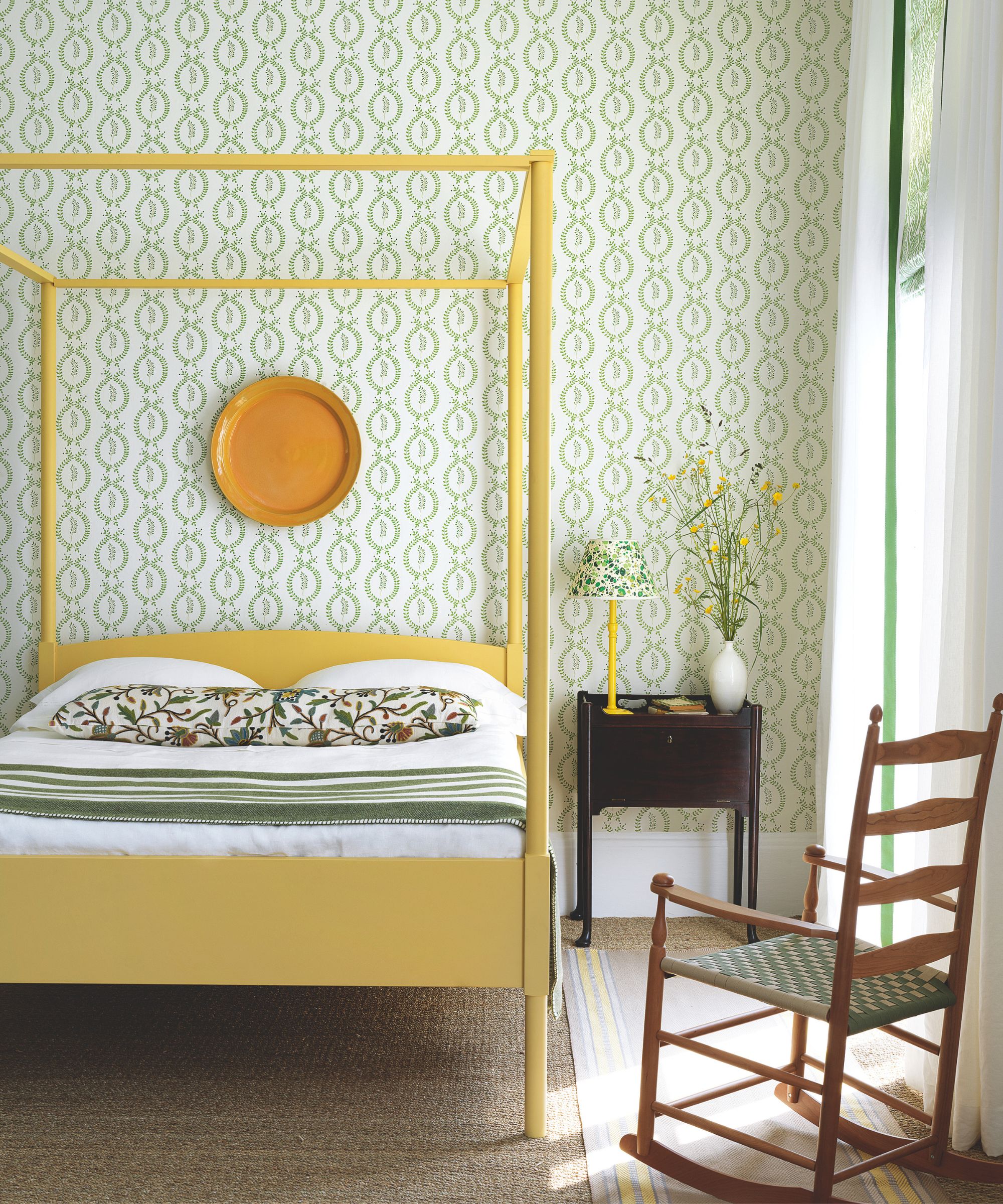 Chambre lumineuse avec papier peint vert et lit à baldaquin jaune, avec une literie blanche, un long coussin à fleurs et une couverture à rayures vertes et blanches, une table de nuit marron et une chaise à bascule en bois.