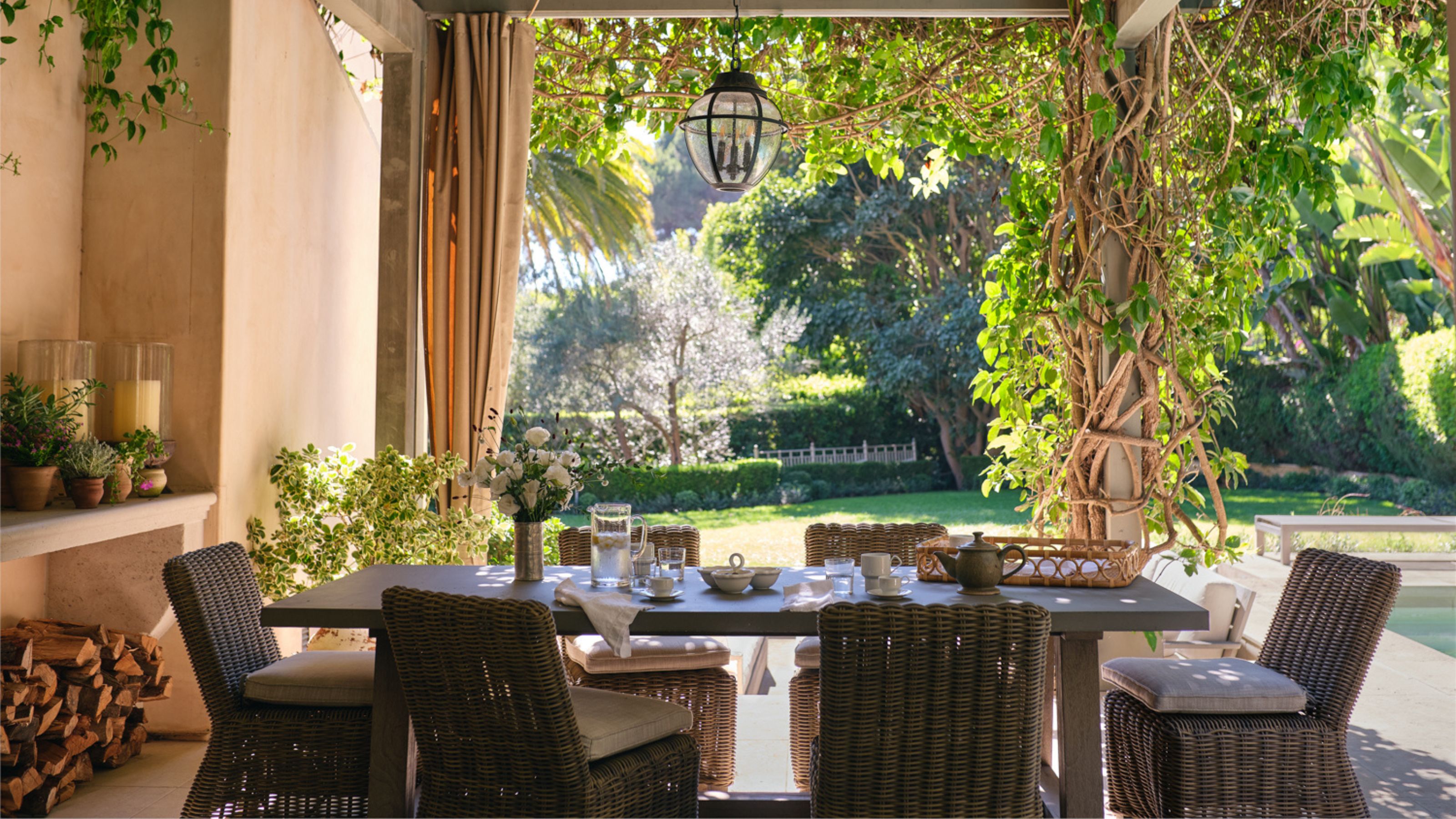 un coin repas extérieur couvert par une pergola avec des chaises en rotin et une table dressée pour le petit-déjeuner