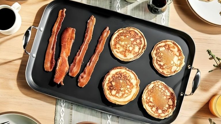 La plaque chauffante brillante à double brûleur de HexClad double l’espace de cuisson, réduit le temps de nettoyage et facilite l’hébergement HexClad Hybrid Griddle with pancakes and bacon on it, on a wooden dining table.