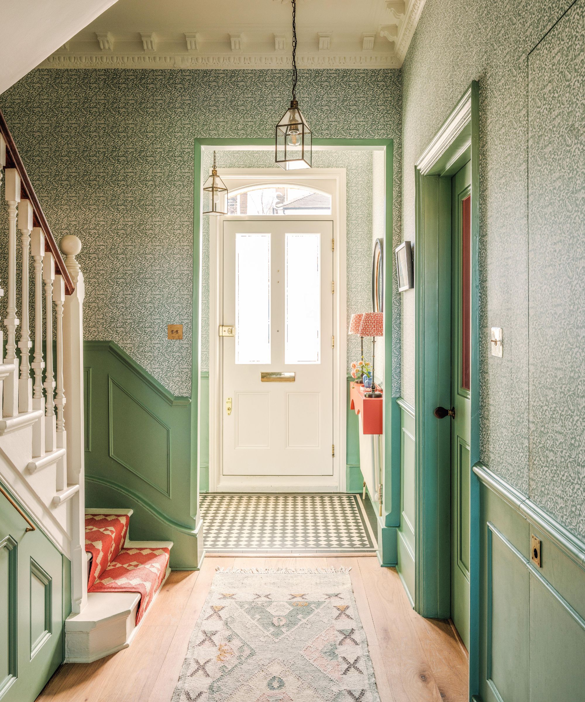 une grande entrée victorienne avec un escalier peint en blanc, une rampe rouge sur les escaliers, du papier peint et des lambris verts sur les murs et un porche avec du carrelage au sol
