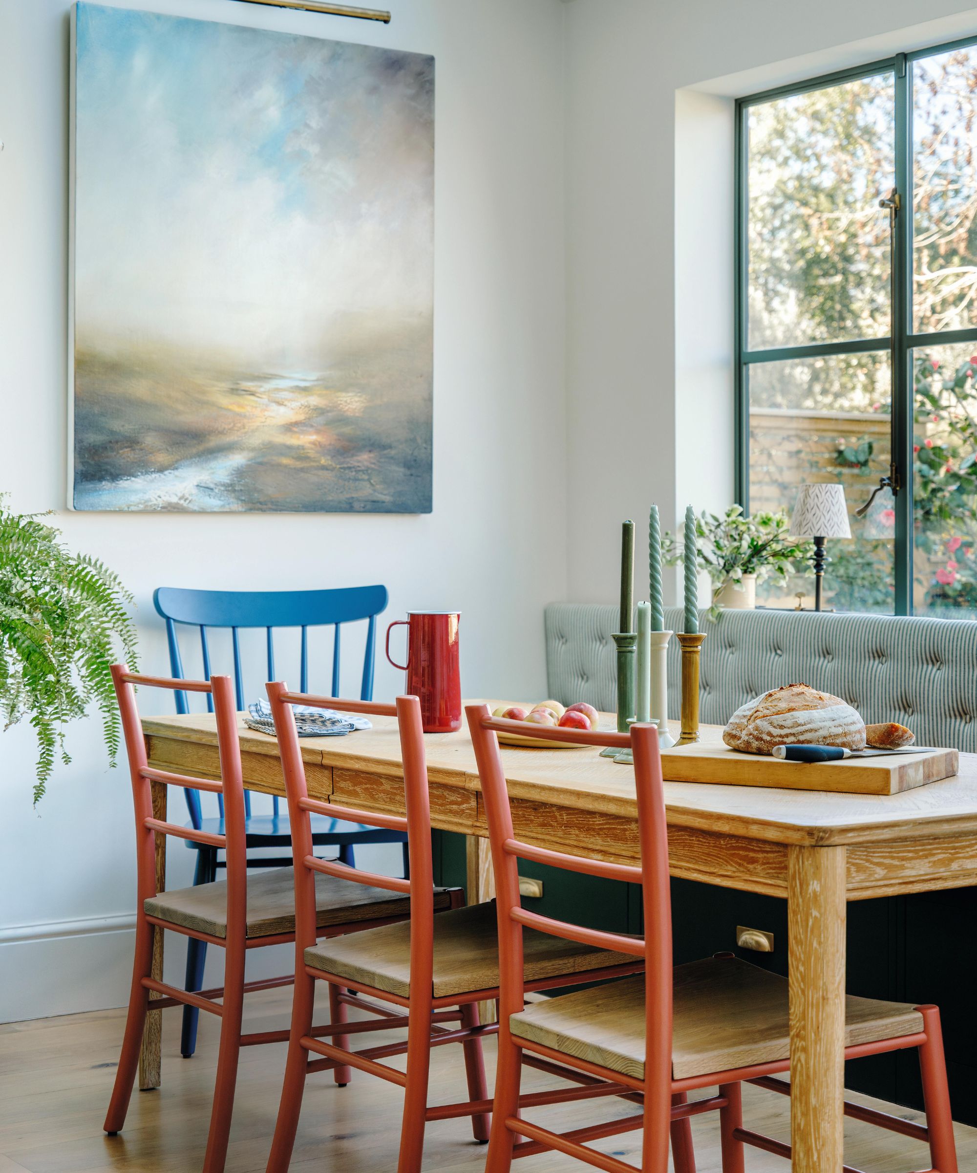 un coin repas avec des murs blancs et une grande fenêtre donnant sur le jardin avec une banquette à rayures vertes construite en dessous. Une table en bois rustique est associée à des chaises rouges