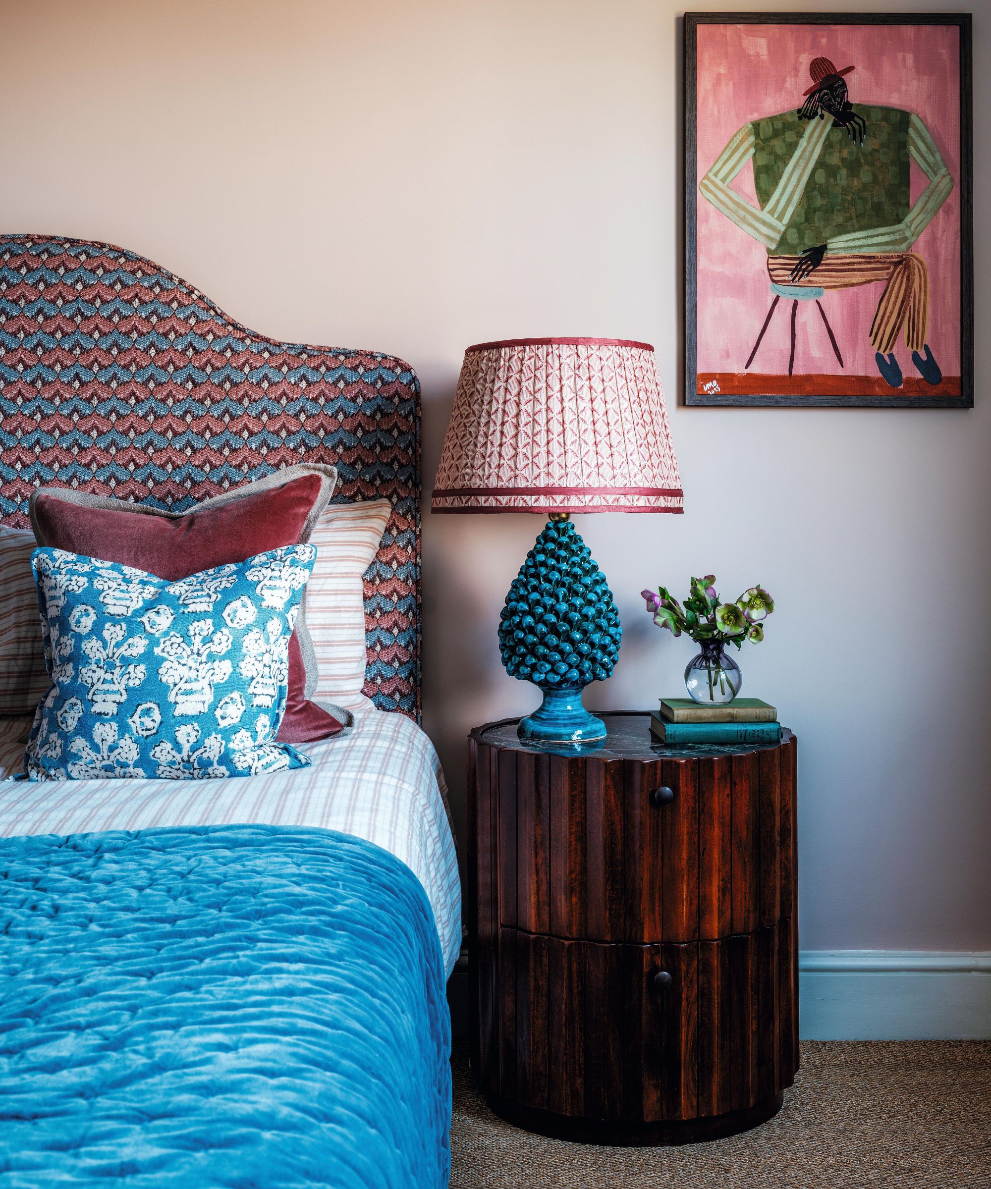 une chambre principale peinte en rose pâle avec un tapis en sisal et une tête de lit à motifs rouges et bleus avec des draps dépareillés assortis et une table de nuit cannelée en bois sombre