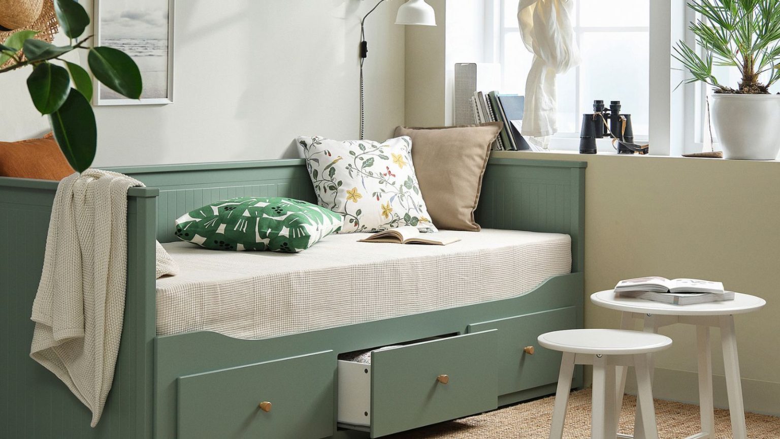 Le lit 3-en-1 le plus vendu d’IKEA vient de bénéficier d’un rafraîchissement chic – apportant l’une des plus grandes tendances de couleurs de 2026 aux petits espaces IKEA grey green HEMNES bed in a living room with floral cushions, large white windows, a pair of white round tables and a jute rug.