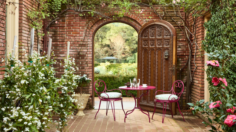 GreenRow vient de lancer une collection enchanteresse avec le jardin botanique de New York – et elle regorge de vaisselle florale nostalgique pour l’hébergement d’été a courtyard with brick walls, a large wooden door, and a red bistro set