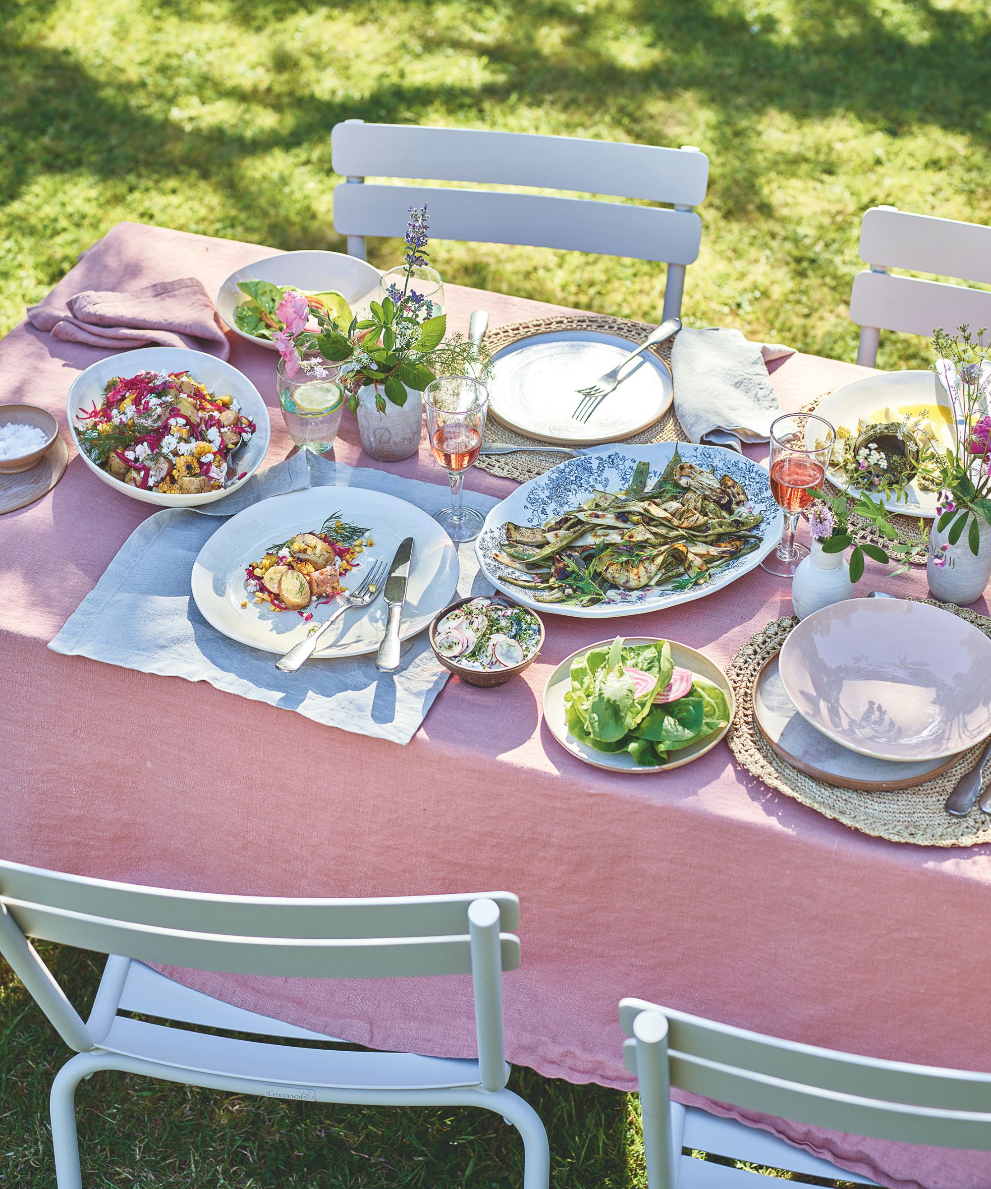 Table à manger extérieure avec chaises de bistro en métal, nappe en lin rose, assiettes de légumes grillés et salades, sets de table en rotin, assiettes blanches, verres à vin et vases à bourgeons remplis de tiges fraîches.