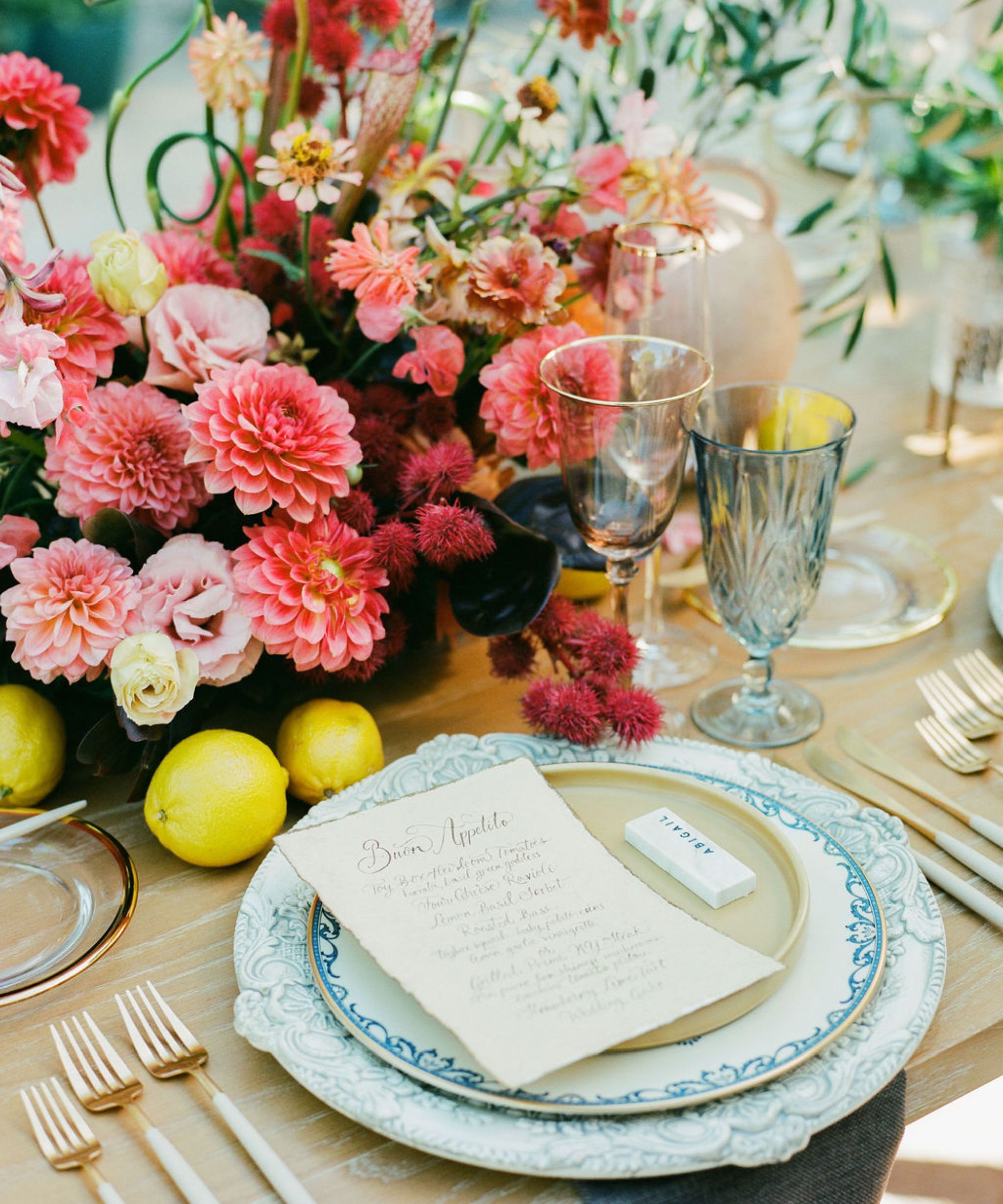 Un décor de table avec des dahlias roses et des fleurs au centre, des assiettes en porcelaine bleue et des couverts dorés, des citrons éparpillés sur la table et de la verrerie bleue.