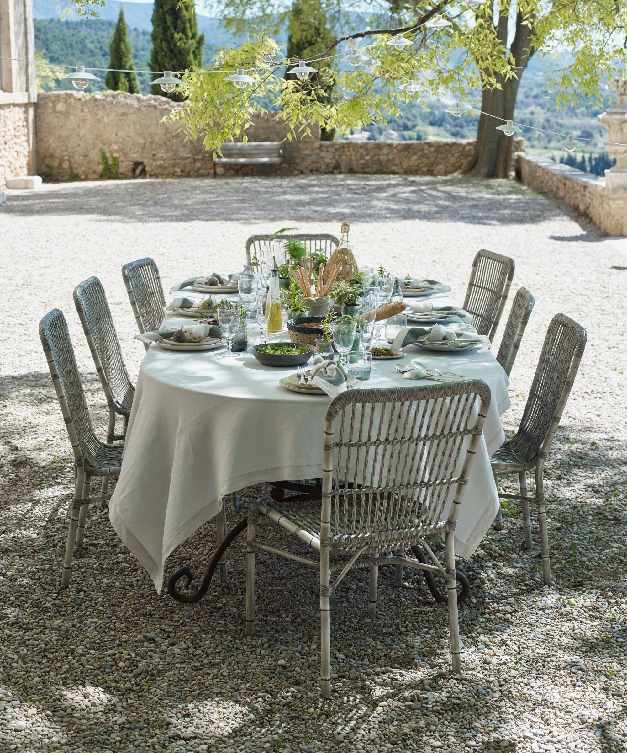 Une salle à manger en plein air dans un jardin méditerranéen avec des arbres, une nappe blanche dessus, des chaises vintage en métal, une table composée d'assiettes vertes et blanches, de verrerie et de fleurs vertes fraîches.