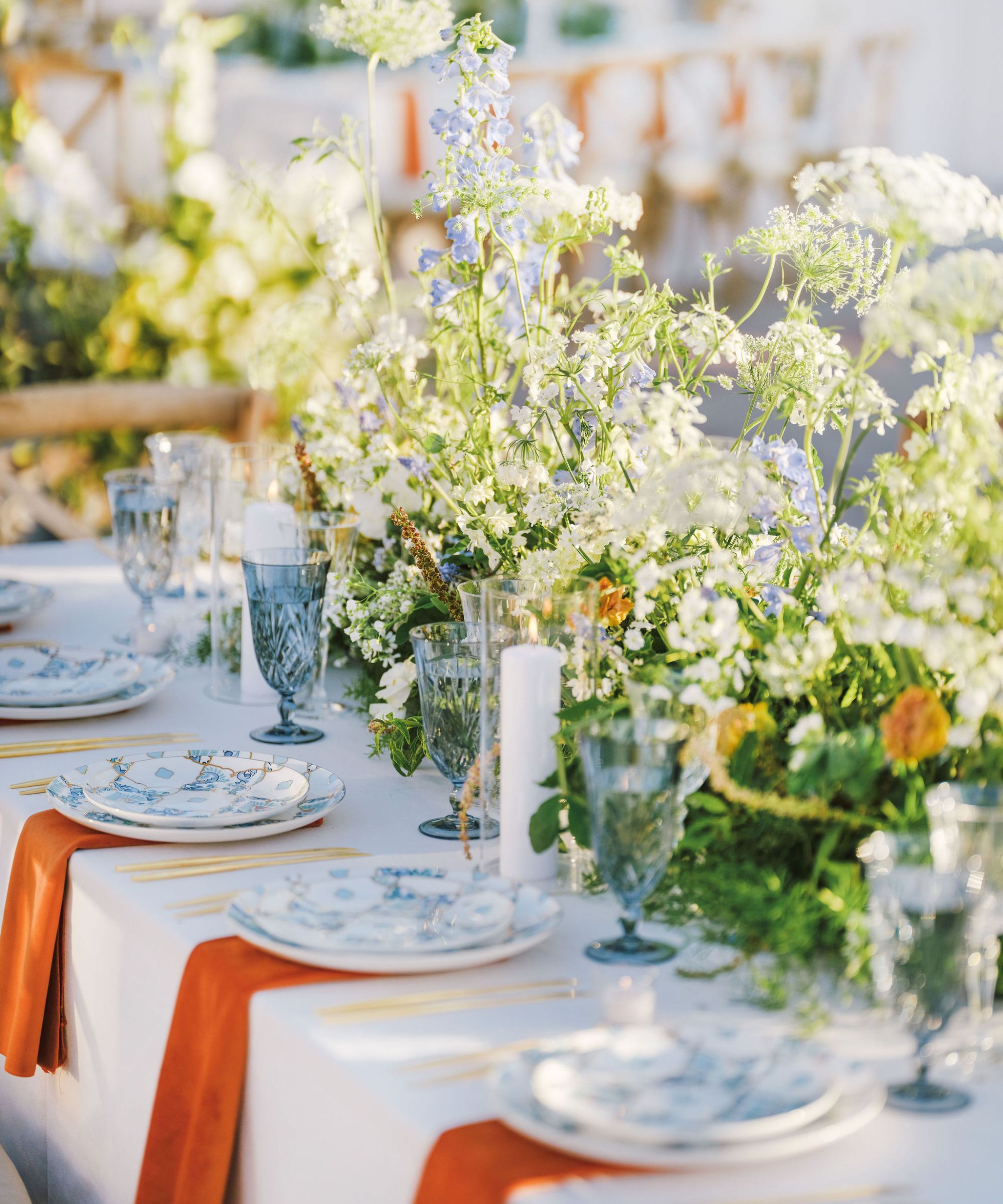Une longue table avec une nappe blanche, des assiettes en porcelaine bleue et blanche, des serviettes orange, des verres bleus et un grand arrangement floral au centre avec de délicates fleurs blanches, violettes et orange.