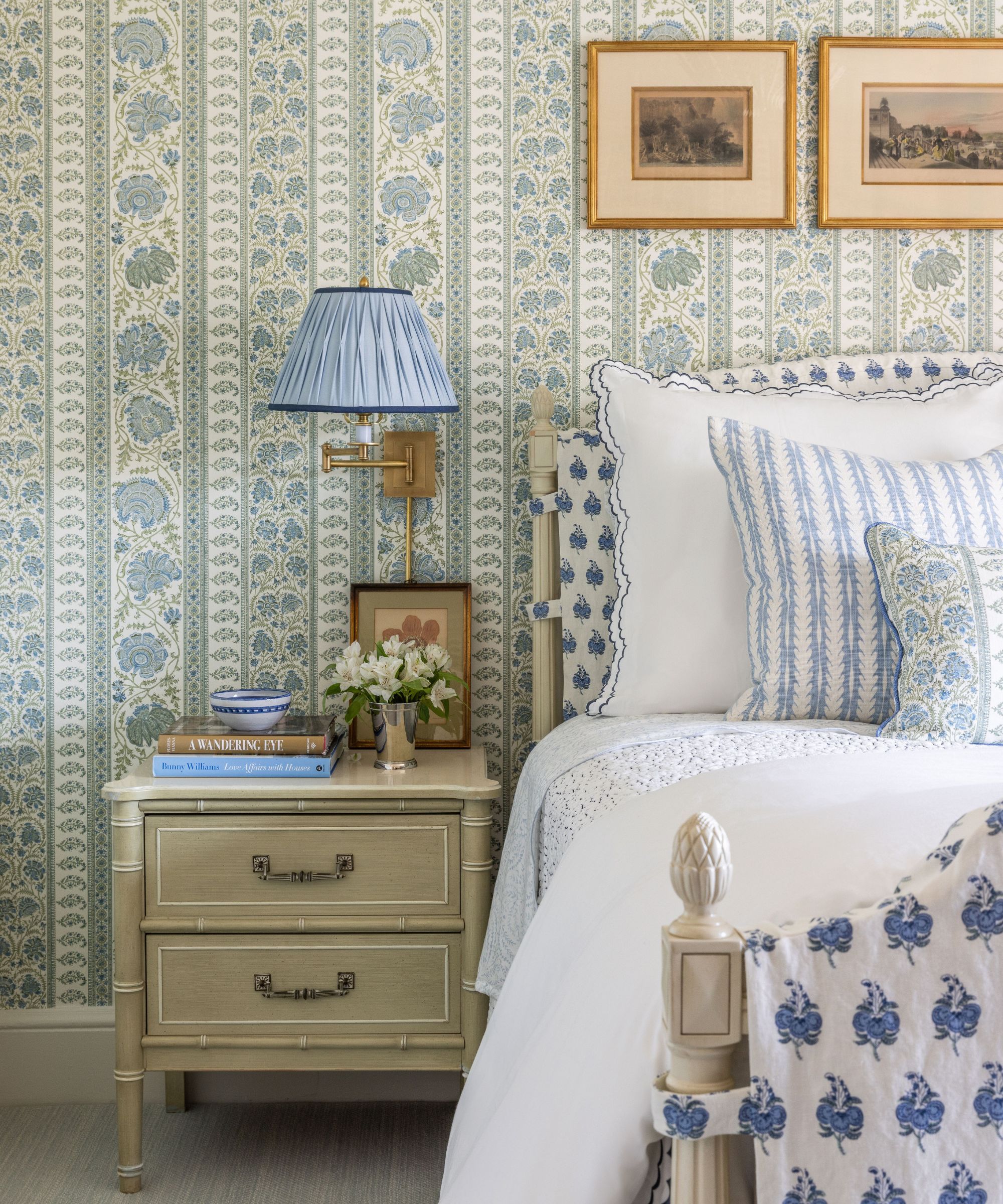 Chambre avec papier peint à rayures cachemire vert et bleu, un lit en bois blanc avec revêtement en tissu, du linge de lit blanc avec des coussins fleuris bleus et une table de nuit en bambou avec des livres et une applique au-dessus.