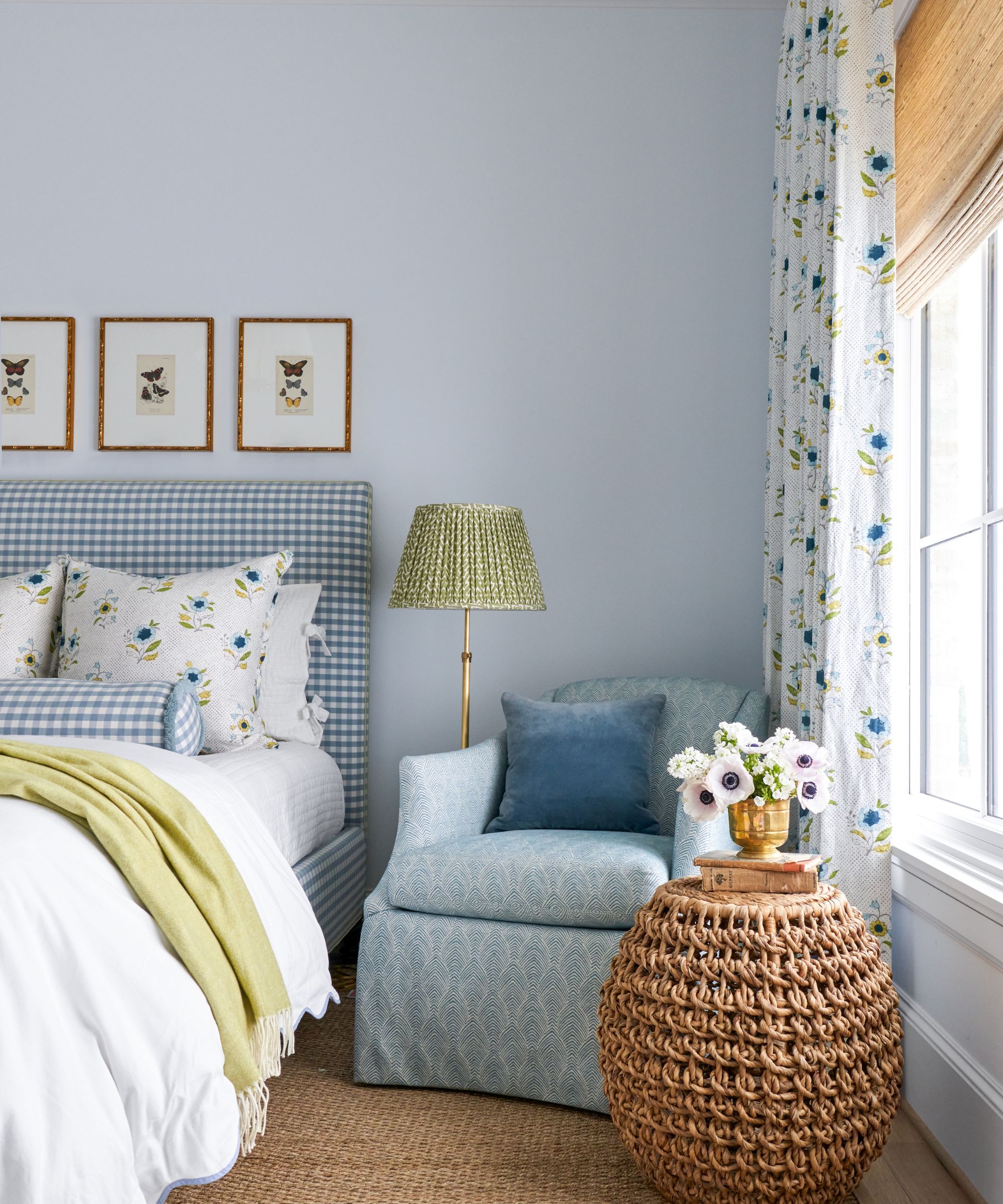 Chambre avec murs peints en bleu pâle, rideaux fleuris et store en bambou, tapis en jute, fauteuil bleu dans le coin avec lampadaire et table d'appoint en osier, et lit rembourré en vichy bleu.