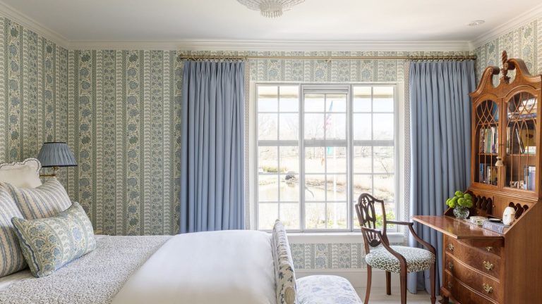 Il est prouvé que cette palette de couleurs pour chambre à coucher 2026 rend votre pièce instantanément plus calme et plus fraîche – et non, vous n’avez pas besoin de repeindre. Bedroom with blue and green floral wallpaper, a large window with blue curtains, a mahogany desk filled with books and a chair underneath, and a white wooden bed with bed linen and floral cushions.