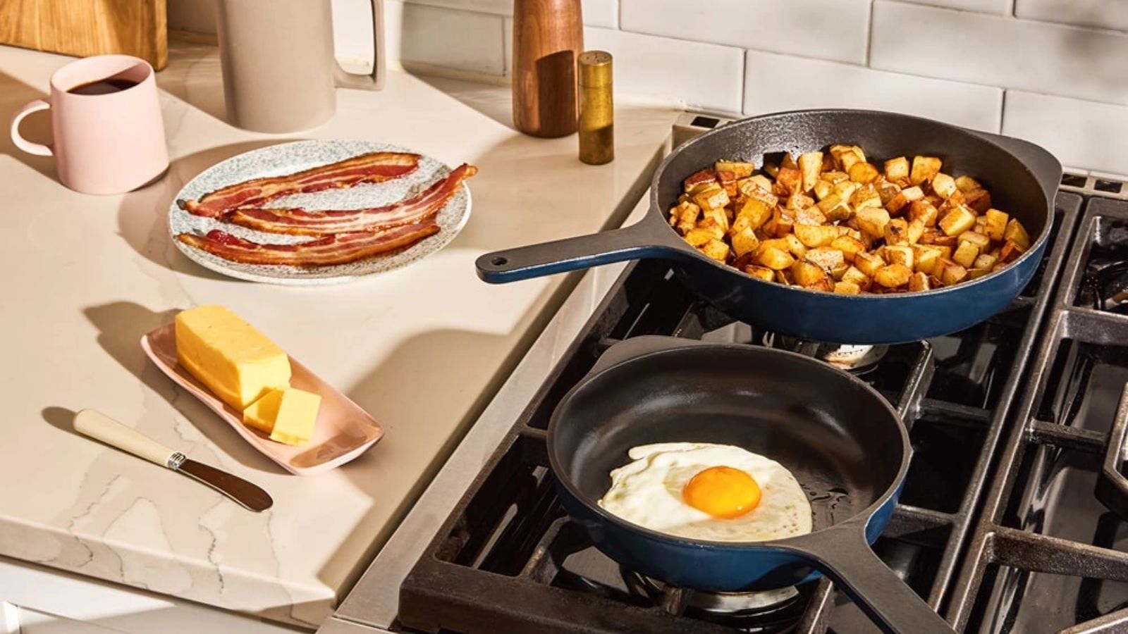 Deux poêles en fonte bleue sur une cuisinière à gaz, une avec des cubes de pommes de terre frites, une avec un œuf au plat. Un morceau de beurre et du bacon frit sur le comptoir à côté d'eux. 
