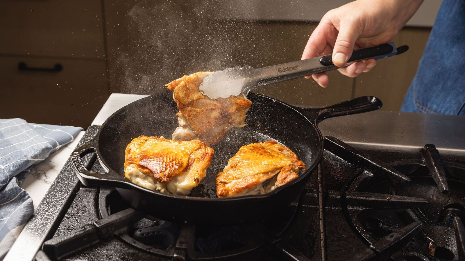 Quelqu'un retourne des cuisses de poulet avec la peau dans une poêle en fonte avec des pinces métalliques. 