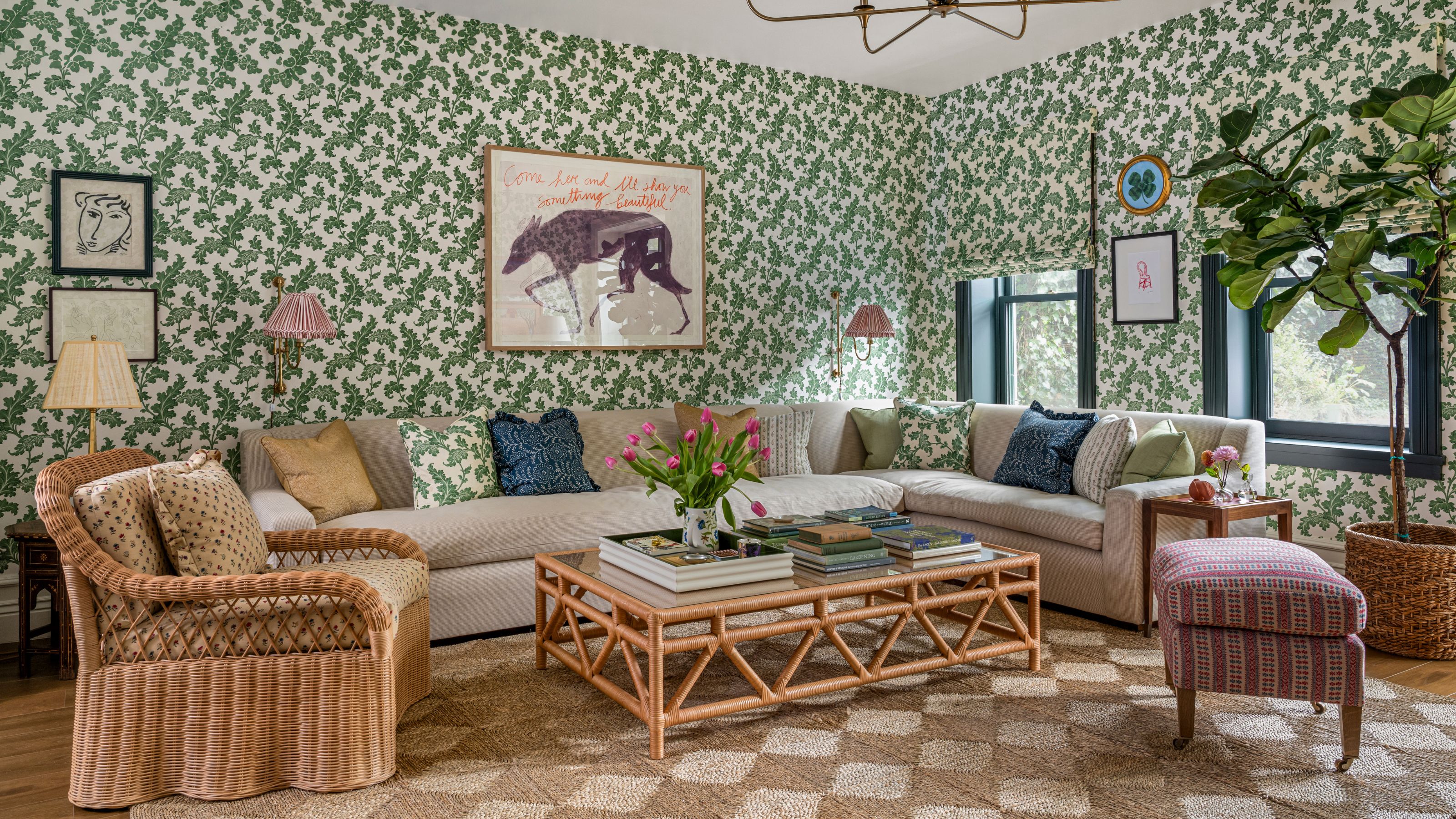 une salle familiale florale verte avec des stores assortis sur deux fenêtres à droite. au centre se trouve un grand canapé sectionnel neutre avec des oreillers floraux, un tapis en jonc de mer sous les pieds, un fauteuil en rotin et une table basse.