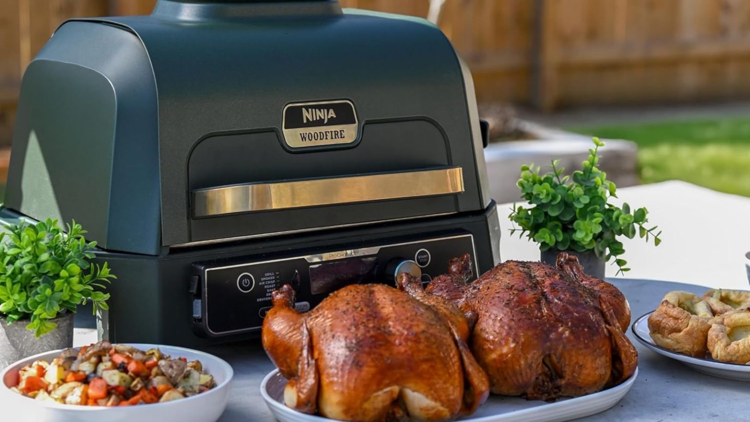 Le gril électrique Ninja’s Genius élimine le stress des barbecues d’été : une cuisson plus rapide, moins de fumée et 100 $ de réduction dès maintenant The blue and gold Ninja Woodfire Pro electric grill on an outdoor table surrounded by fried chickens, sides, and potatoes