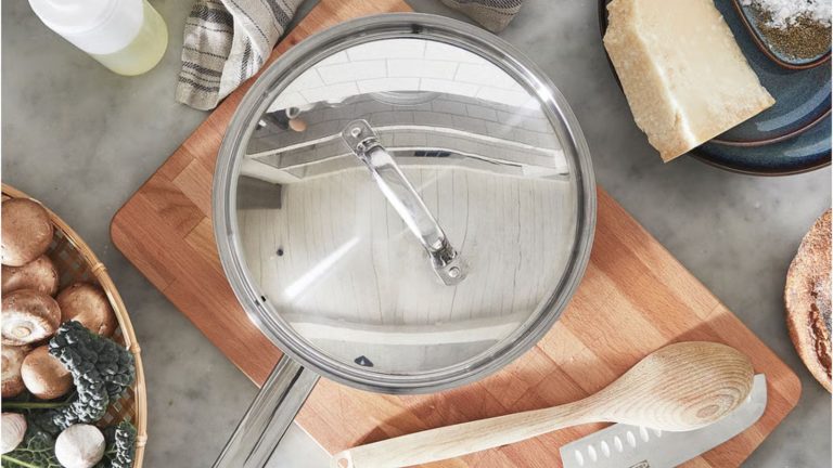A stainless steel saut&eacute; pan with a long handle and matching lid on a wooden cutting board, viewed from above