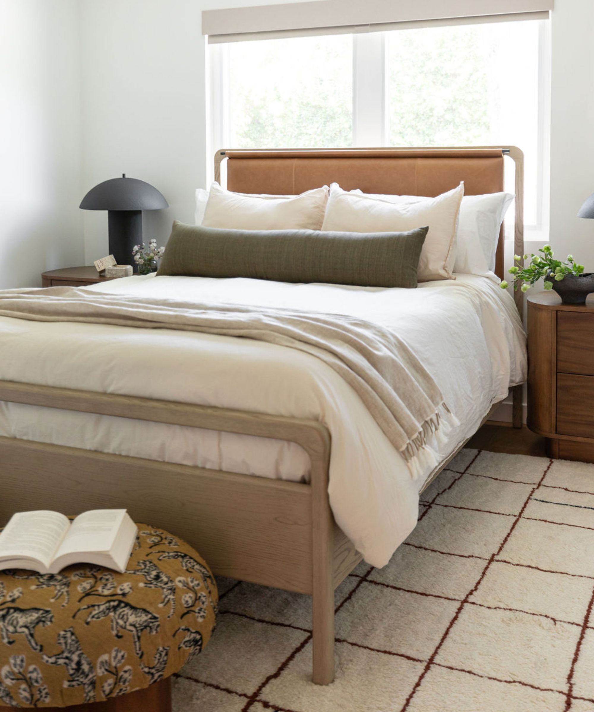 Chambre neutre avec un lit en bois avec une tête de lit en cuir, des draps en lin et un oreiller lombaire marron foncé, un tapis géométrique crème, un pouf rembourré jaune moutarde et une lampe de table ronde noire.