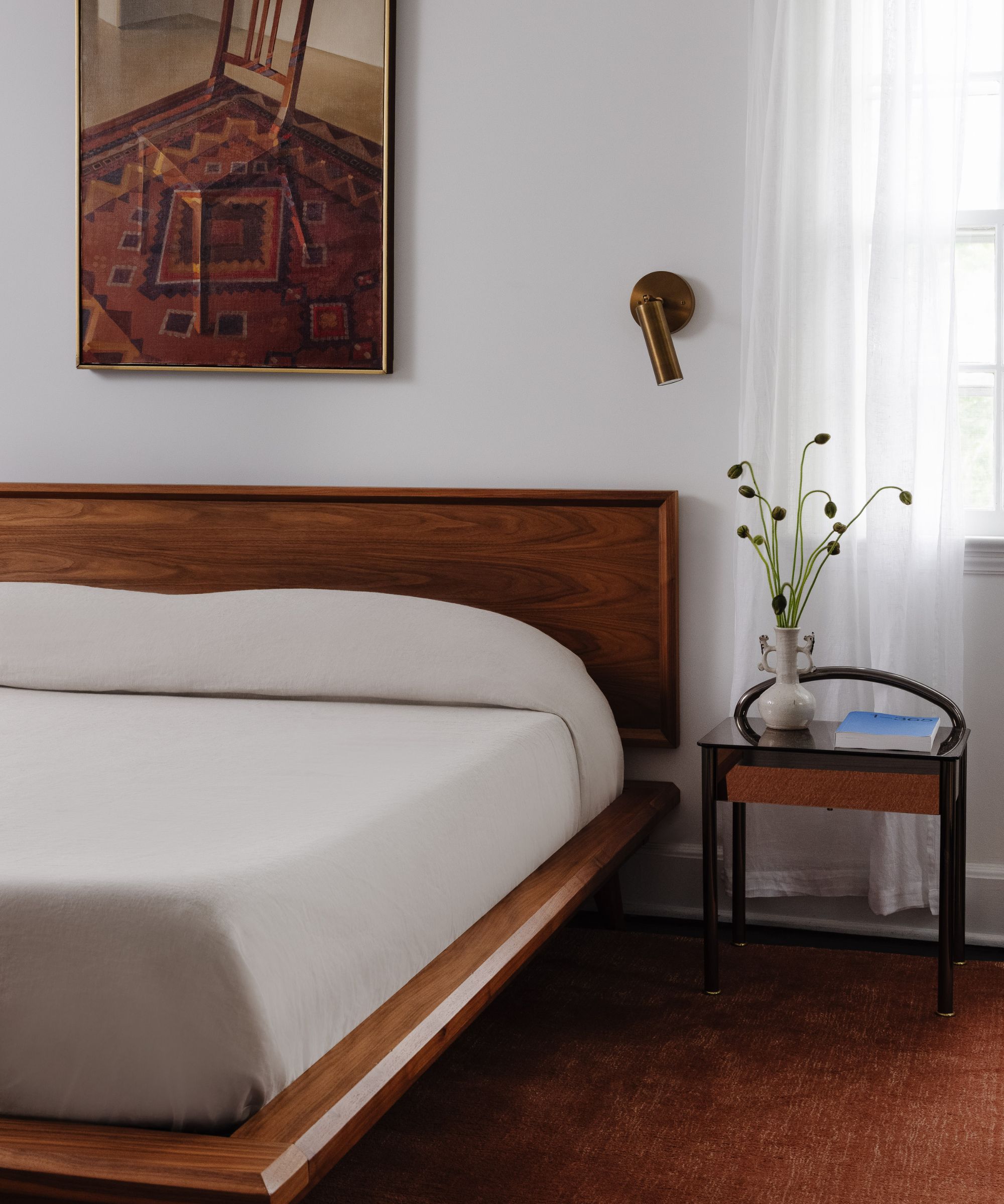 Chambre avec un lit bas en bois du milieu du siècle, une literie en lin neutre, une applique murale dorée, une nature morte représentant une chaise, un tapis rouge foncé et une table de nuit en bois avec un vase de fleurs.