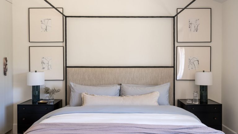 Comment superposer votre lit comme un designer cet été – et pourquoi c’est en fait le simple secret d’une nuit de sommeil plus fraîche Bedroom with a black metal four-poster bed, lilac linen bedding and a neutral lumbar pillow, four framed prints on the wall in black frames, and black nightstands on either side with table lamps.