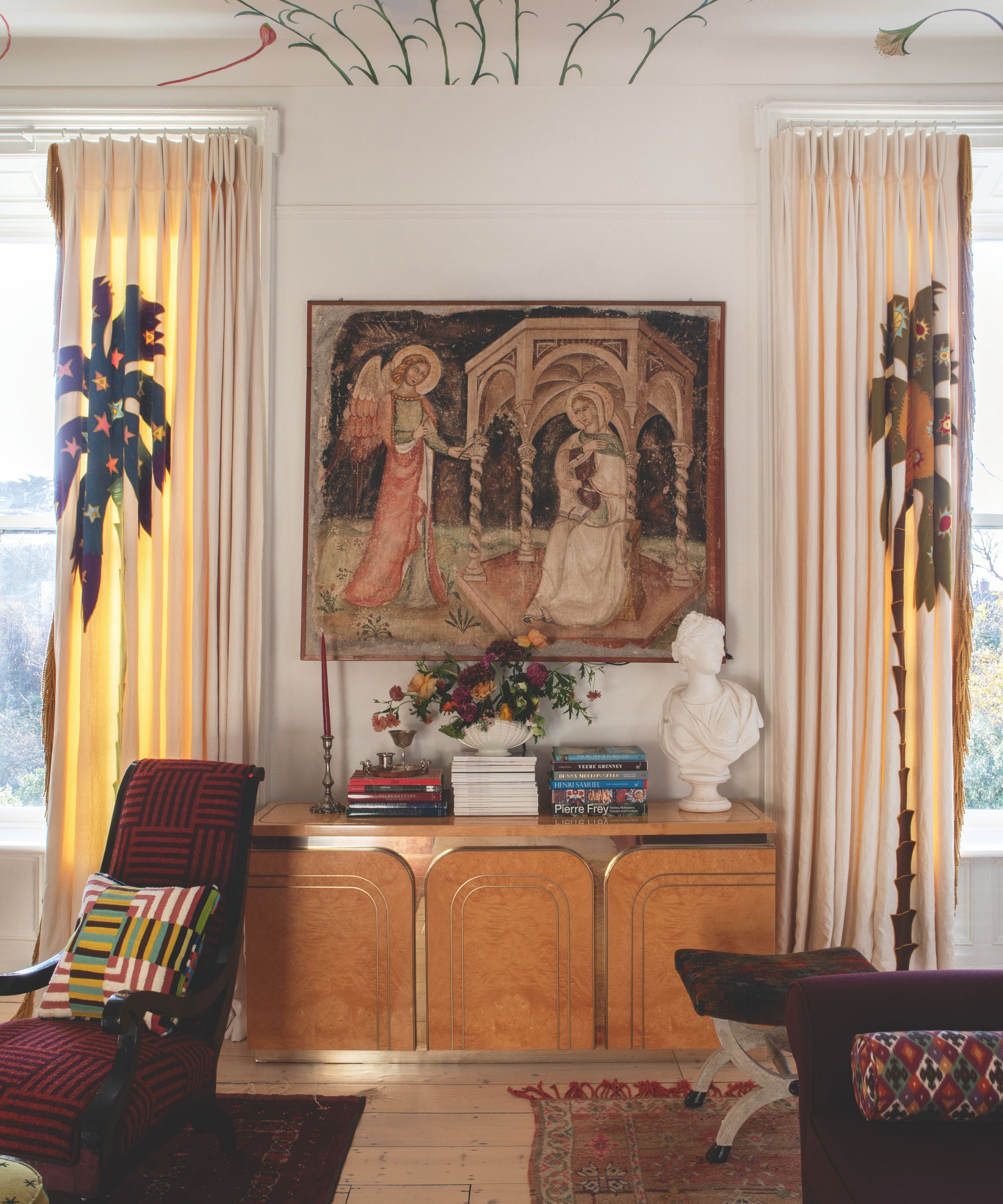Salon avec fresque murale au plafond, console en bois surmontée d'une tapisserie ancienne, buste classique et livres empilés sur la console, fauteuil rouge avec coussin géométrique et repose-pieds.