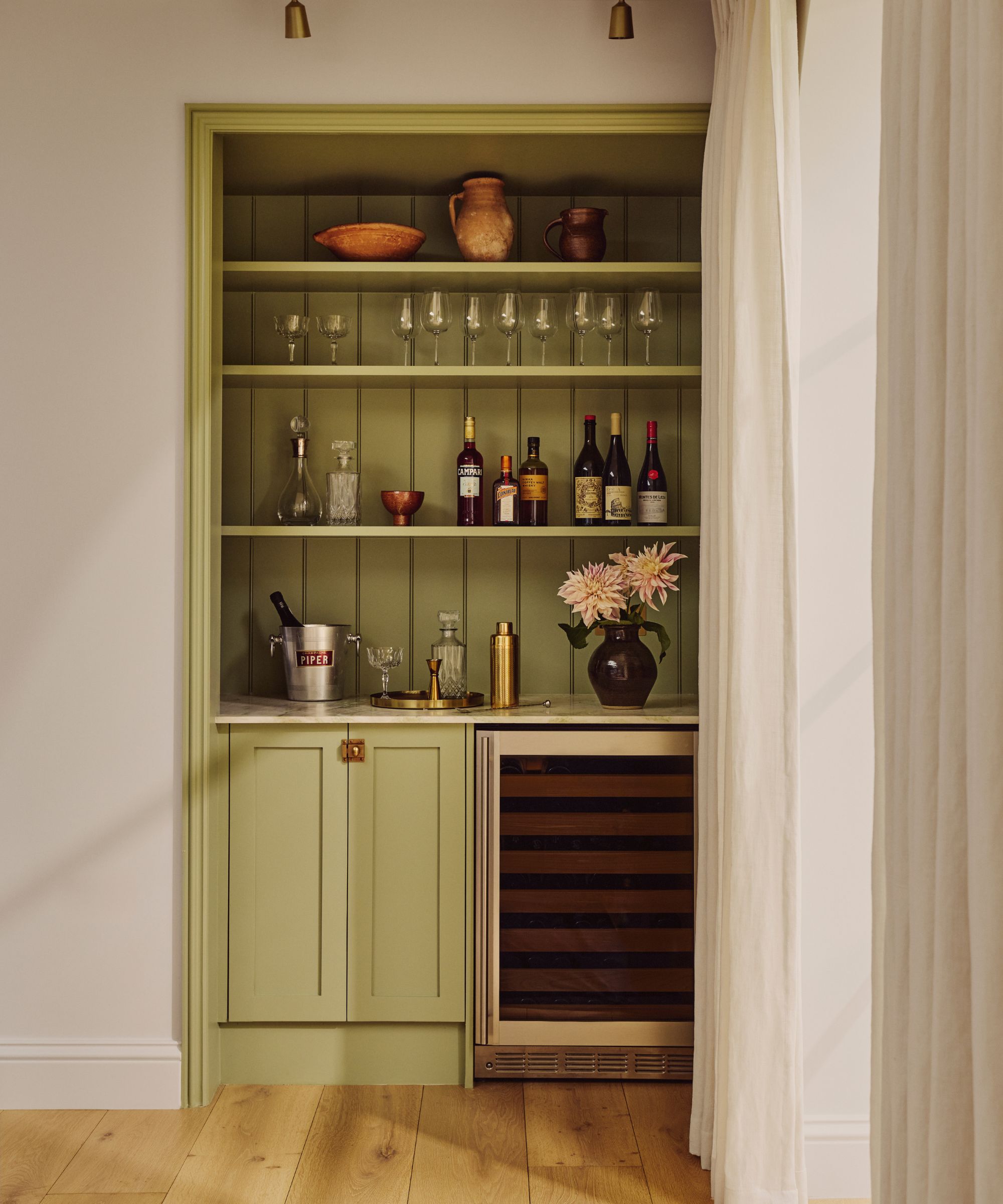 Bar à la maison construit dans un coin, peint en vert, avec des étagères contenant des verres et des bouteilles d'alcool, un shaker et un seau à glace, ainsi qu'un vase de fleurs, avec un mini-réfrigérateur et un placard en dessous.