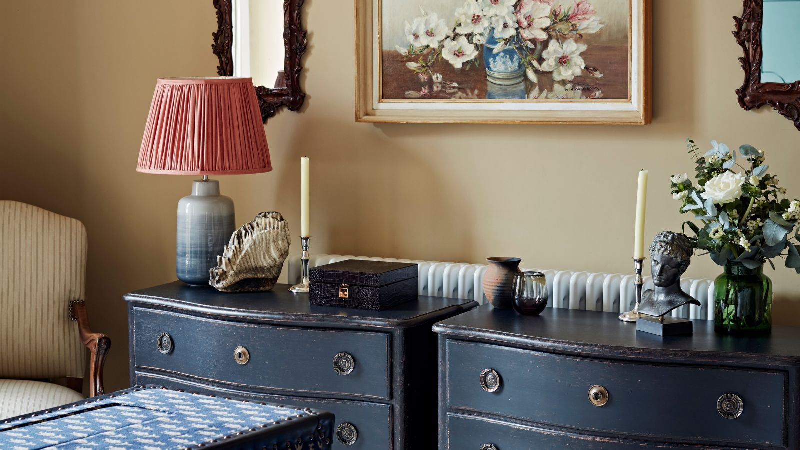 Deux commodes bleu marine foncé avec bougies, fleurs et lampes, contre un mur beige. Ci-dessus se trouvent une nature morte florale et deux miroirs en bois, avec un coffre à motifs et une chaise rembourrée à rayures.