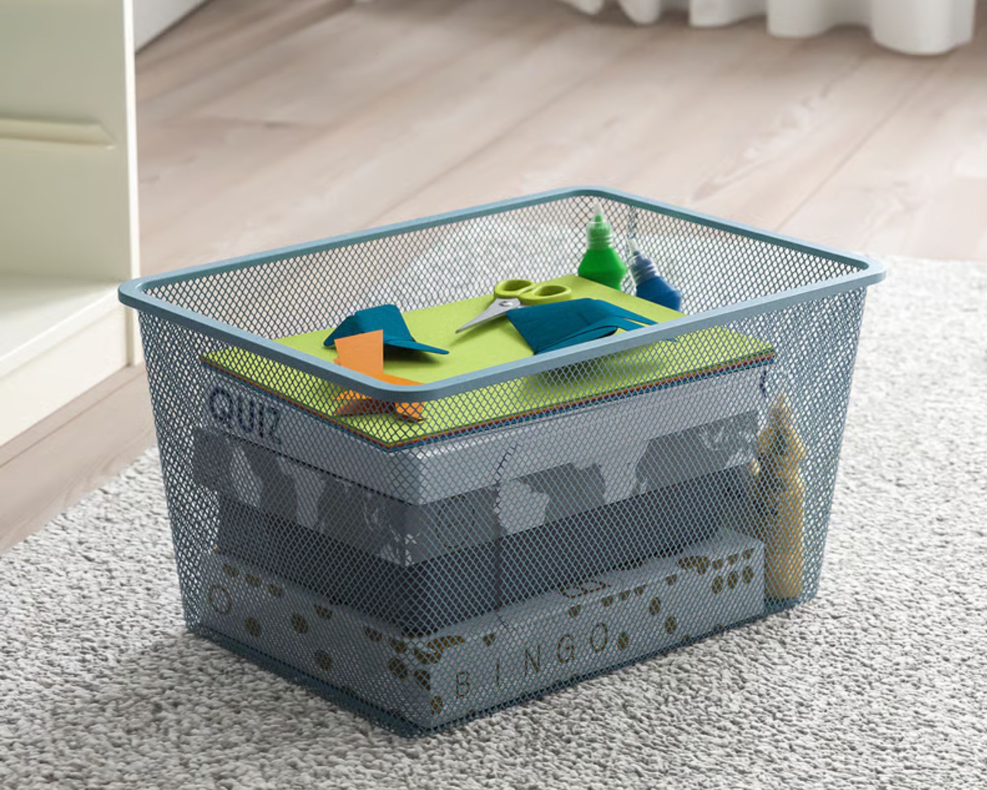 Une boîte de rangement IKEA TROFAST Mesh bleu pâle remplie de jeux, d'origami et de ciseaux, représentée sur un tapis couleur blé sur un parquet en bois clair.