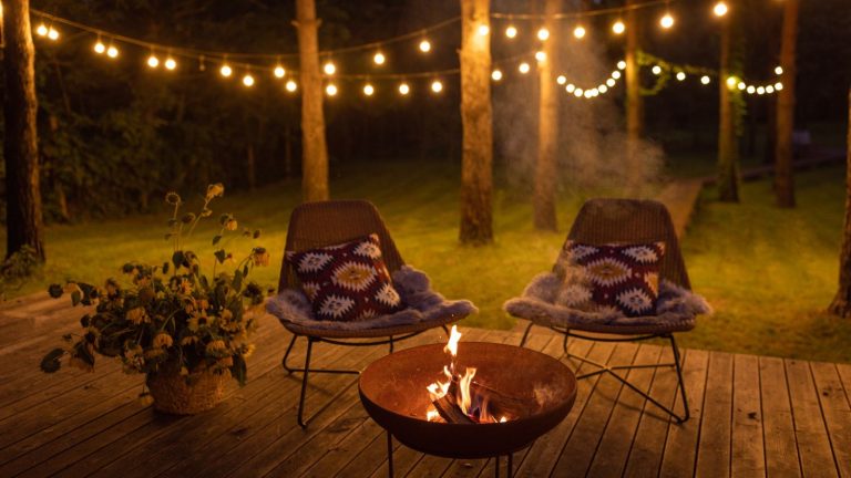Comment utiliser la cendre de bois dans le jardin – Cela pourrait être le booster de sol gratuit que vous négligez A fire pit on a deck surrounded by two comfortable chairs at night, with the garden lit up