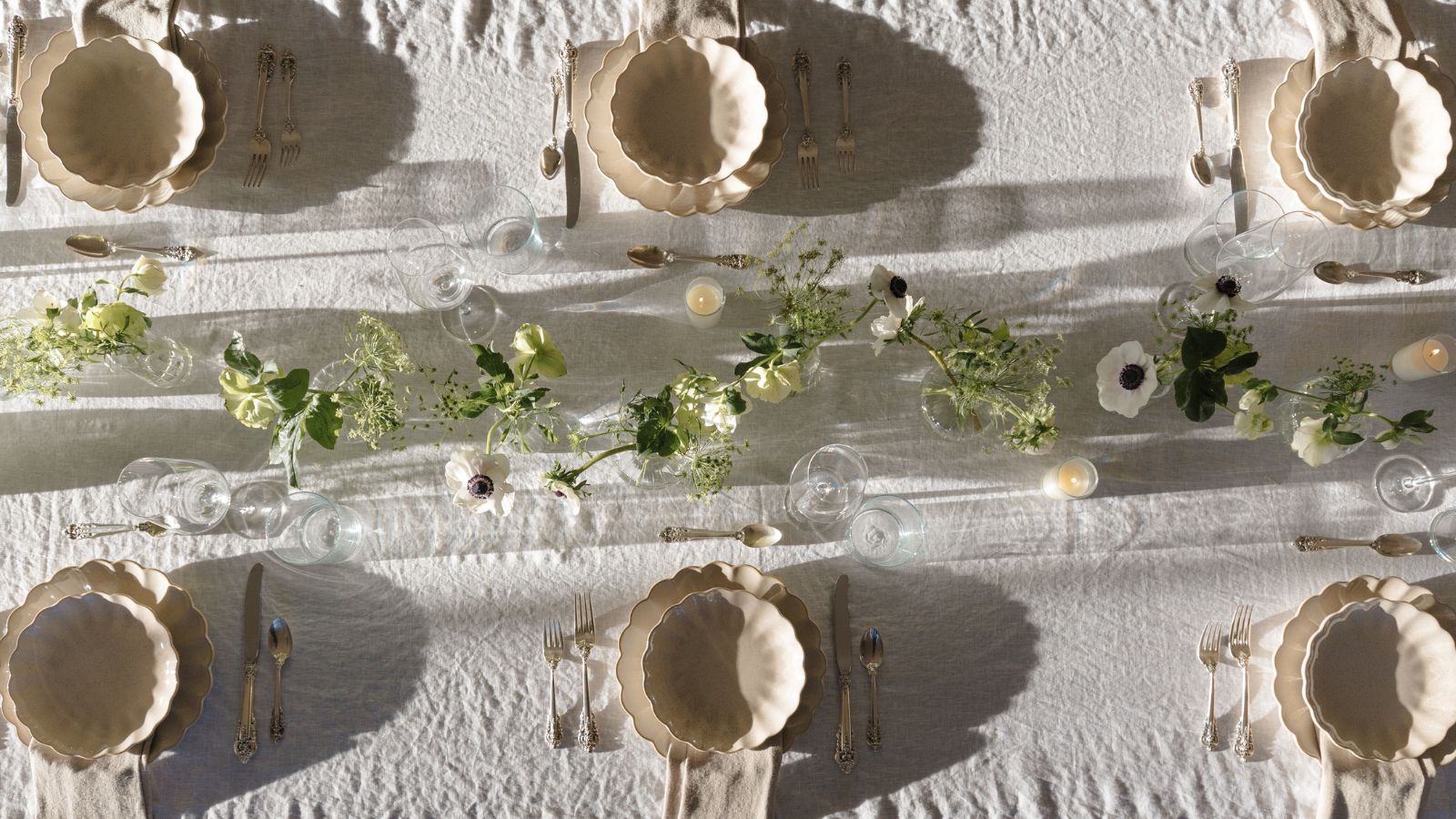Table d'extérieur en lin blanc, assiettes et bols festonnés crème, guirlande florale au centre et bougies allumées