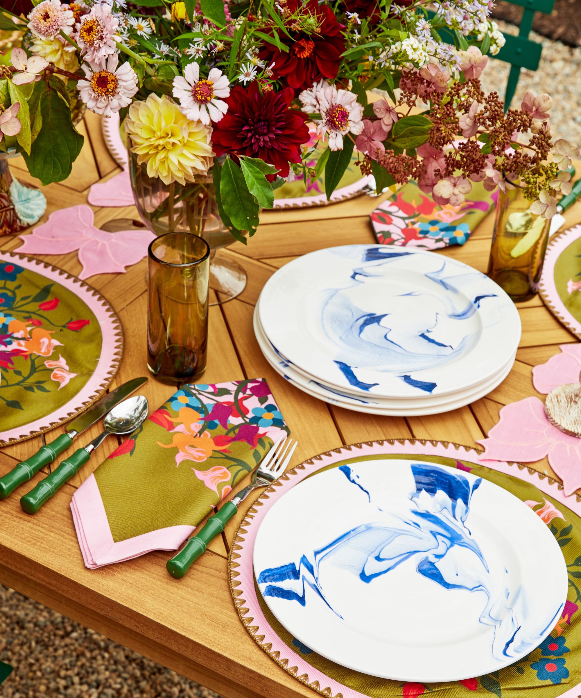 Table en bois avec sets de table colorés, couverts en bambou vert, assiettes blanches et bleues, serviettes fleuries et verres ambrés avec un grand arrangement floral au centre.
