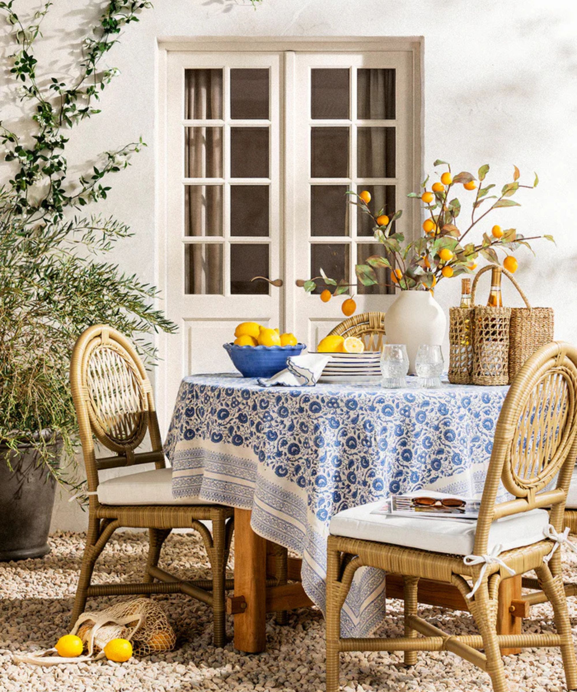 Salle à manger estivale en plein air aménagée avec une nappe bleue et blanche, des chaises en rotin, un bol de citrons, des piles d'assiettes, un vase avec des fleurs d'oranger, un casier à vin en rotin et des citrons au sol.