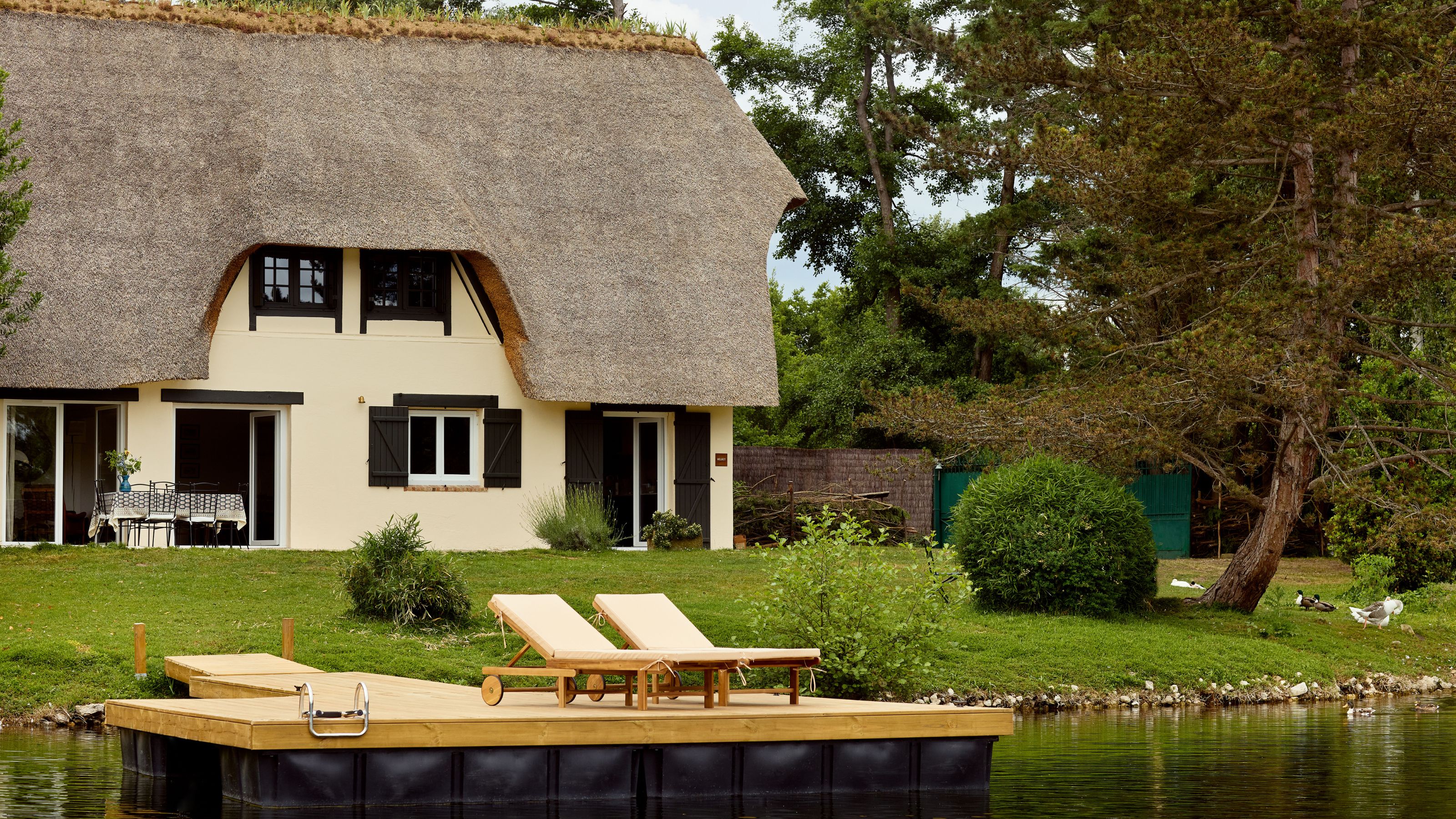 une chaumière au bord d'un étang en Normandie, France entourée de verdure, d'arbres, de canards avec une terrasse pour bronzer
