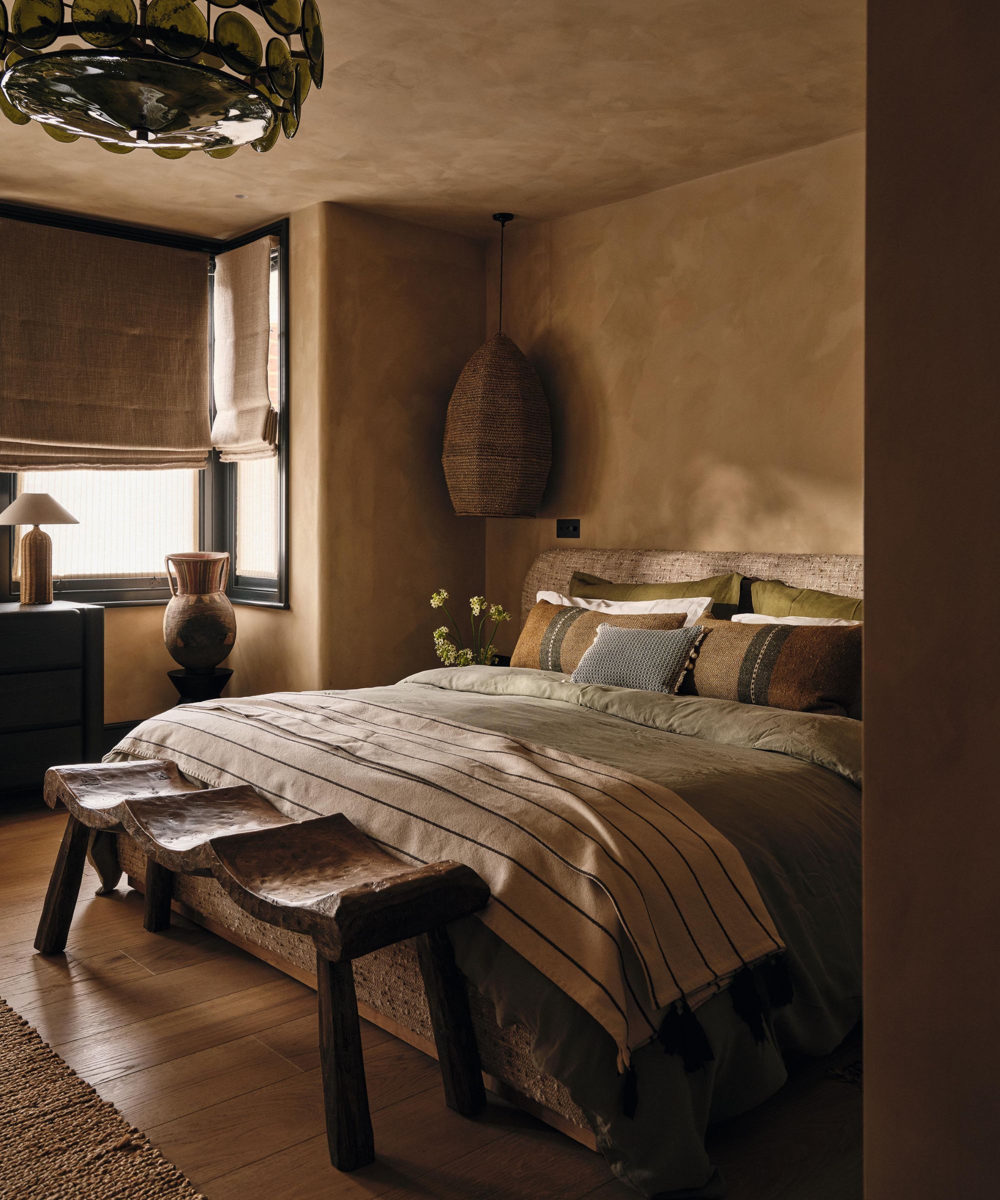 une chambre beige foncé blanchie à la chaux avec un lit en lin, des meubles rustiques en bois, un lustre en verre vert et des pièces en rotin