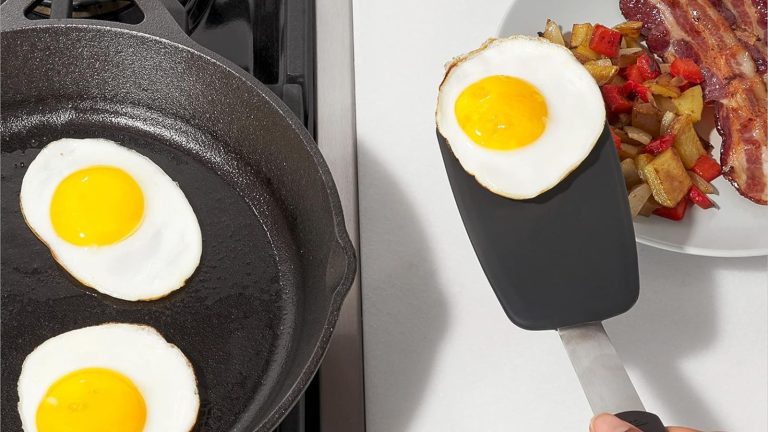 Someone lifting a fried egg out of a cast iron pan and transferring it to a plate with a black spatula