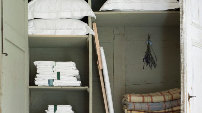 A pale green linen closet with pillows in white pillowcases, folded white sheets, folded gingham blankets, an ironing board, and a sprig of lavender hung up with a blue ribbon.