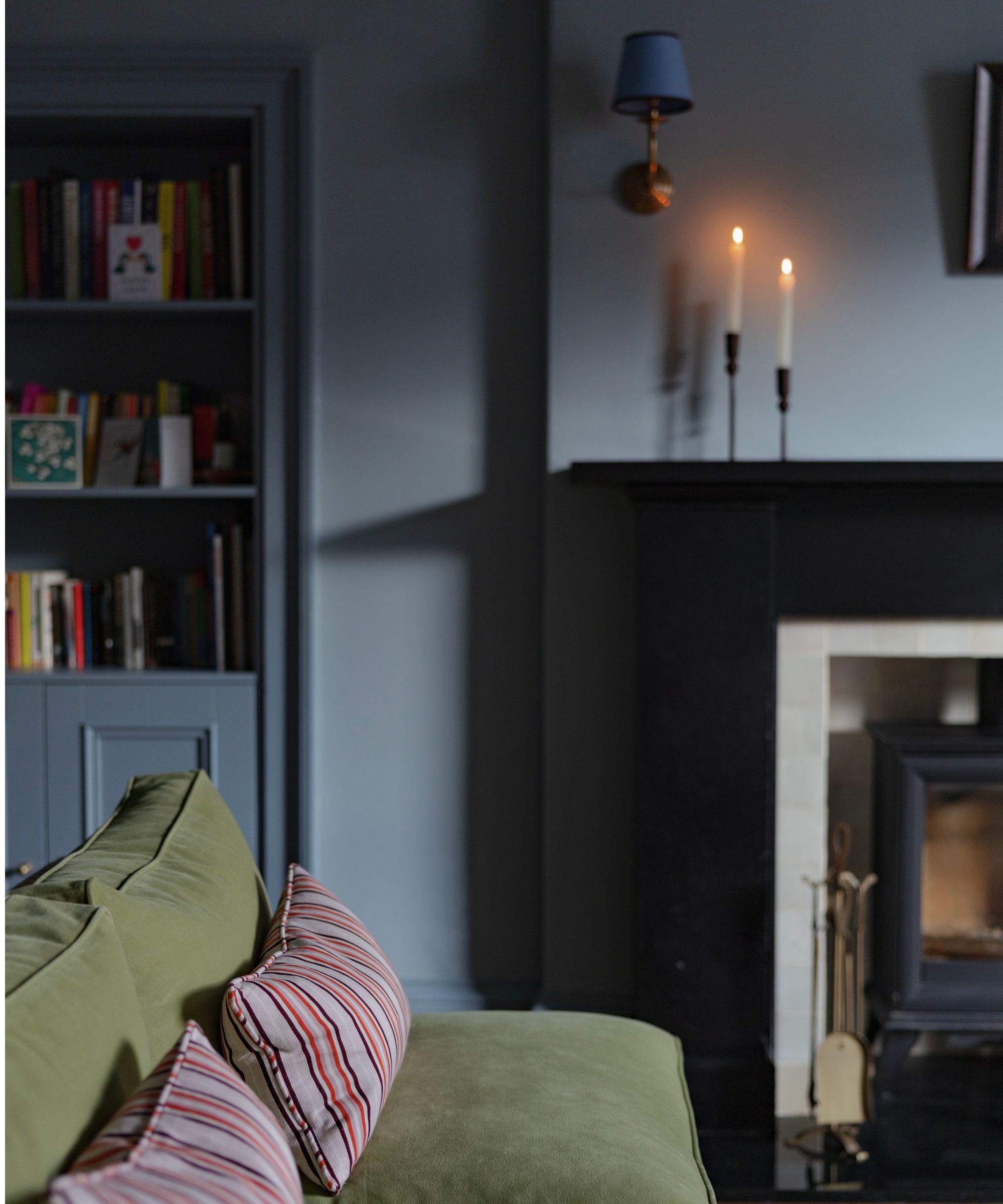 Salon avec des murs bleu-gris profonds, des étagères intégrées, un canapé vert avec des coussins rayés et une cheminée noire éclairée aux chandelles.