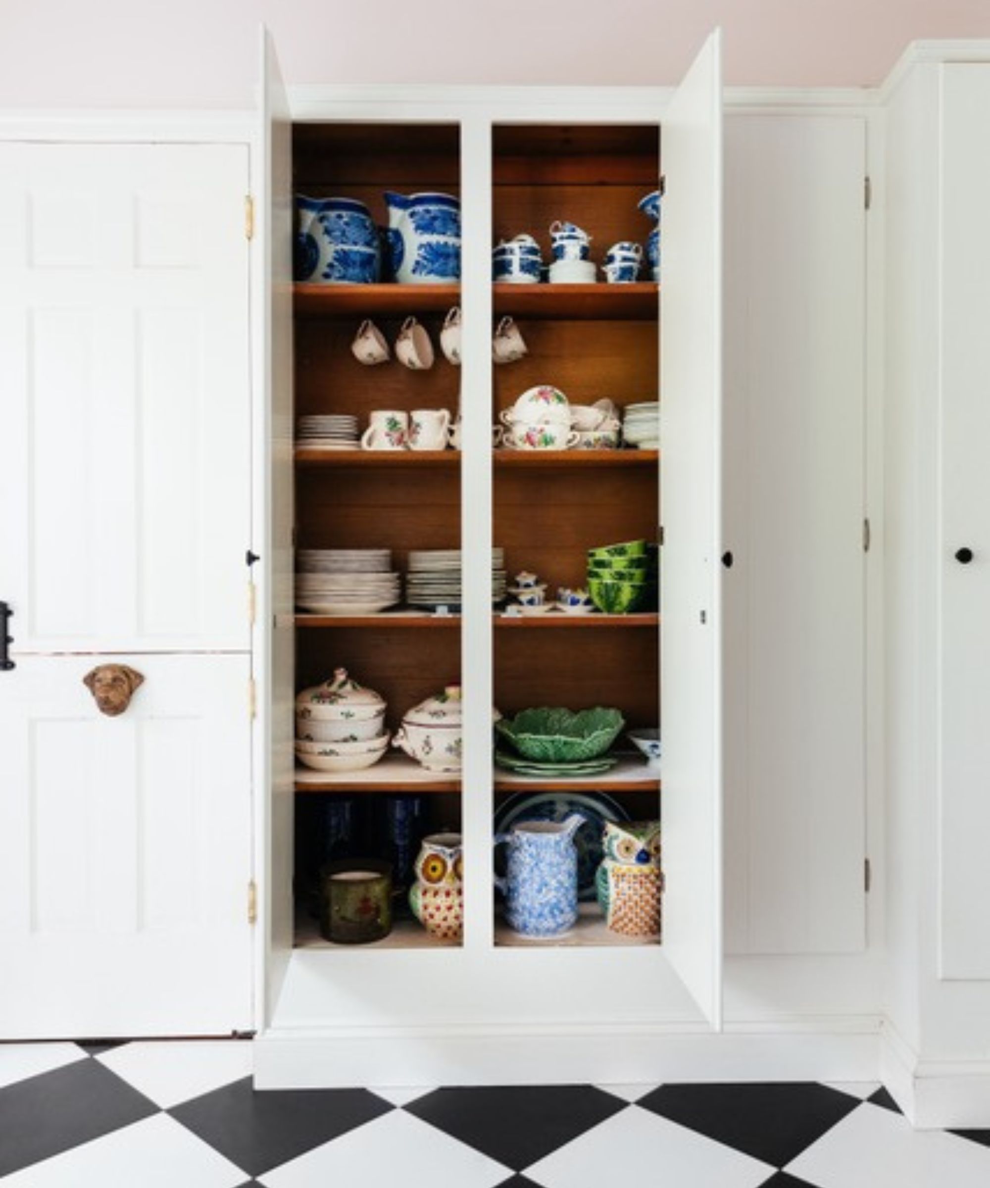 Armoire de cuisine intégrée peinte en blanc avec un plafond rose pâle, présentant des céramiques et de la vaisselle colorées sur des étagères en bois chaleureux au-dessus d'un sol en damier noir et blanc.