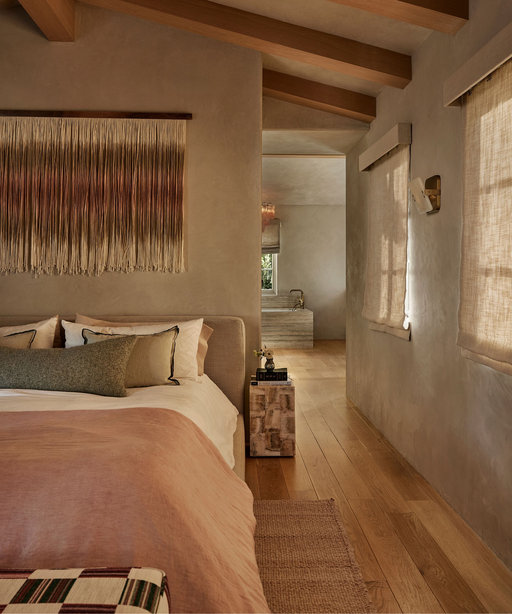 Chambre avec murs en plâtre taupe chaud, poutres apparentes au plafond, stores romains en lin et literie neutre en couches, créant une retraite calme et organique avec des textures naturelles.