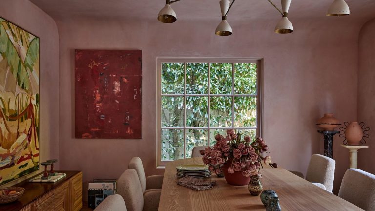 Blush-pink plaster walls, a long oak dining table, sculptural lighting, contemporary art, and lush garden views create a warm, layered dining room with modern organic style.