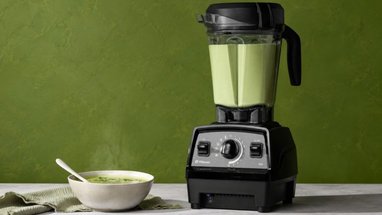 A Vitamix propel series 750 blender with green soup inside, against a green background. There is a small white bowl of green soup beside it.