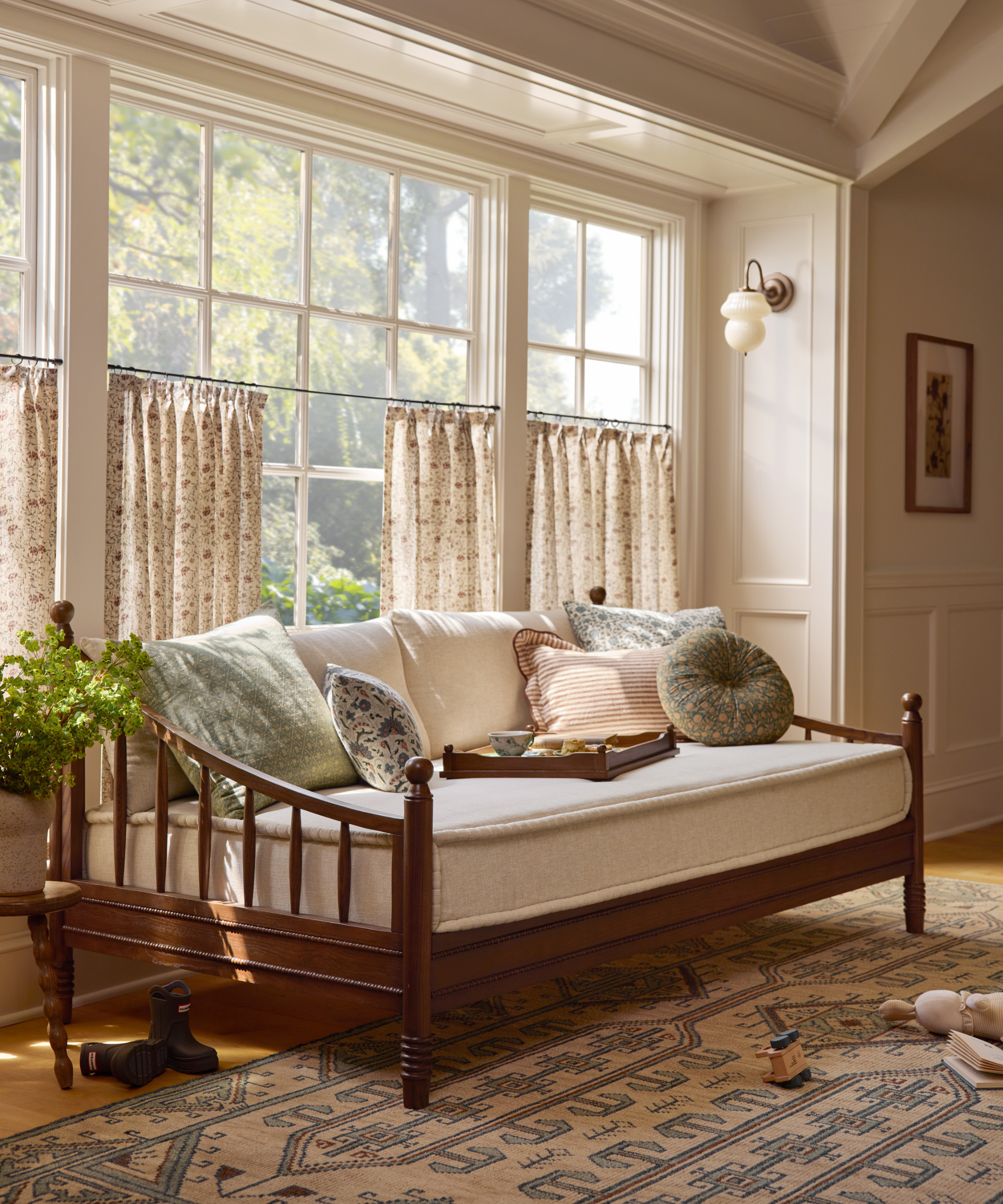 un lit de repos en bois avec un coussin de siège beige, de nombreux oreillers, un tapis géométrique et des rideaux de café le long des fenêtres