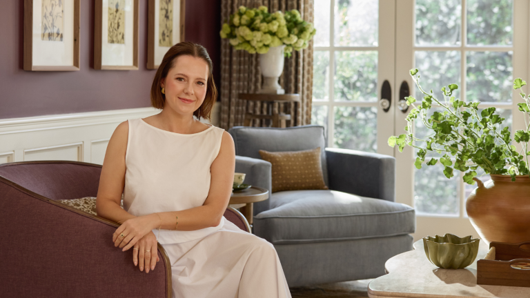 a woman sitting on a maroon chair in a living room with several plants and a blue accent chair in the background