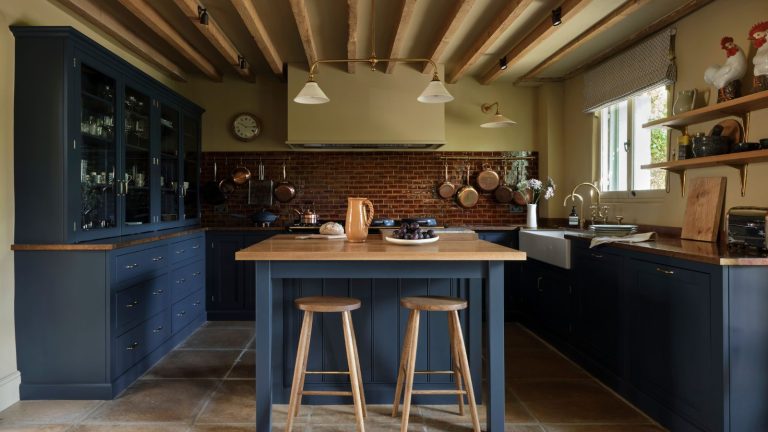 Dark blue kitchen with navy cabinetry, oak countertops, copper cookware, brass fixtures, and exposed timber beams, creating a warm and cocooning feel.