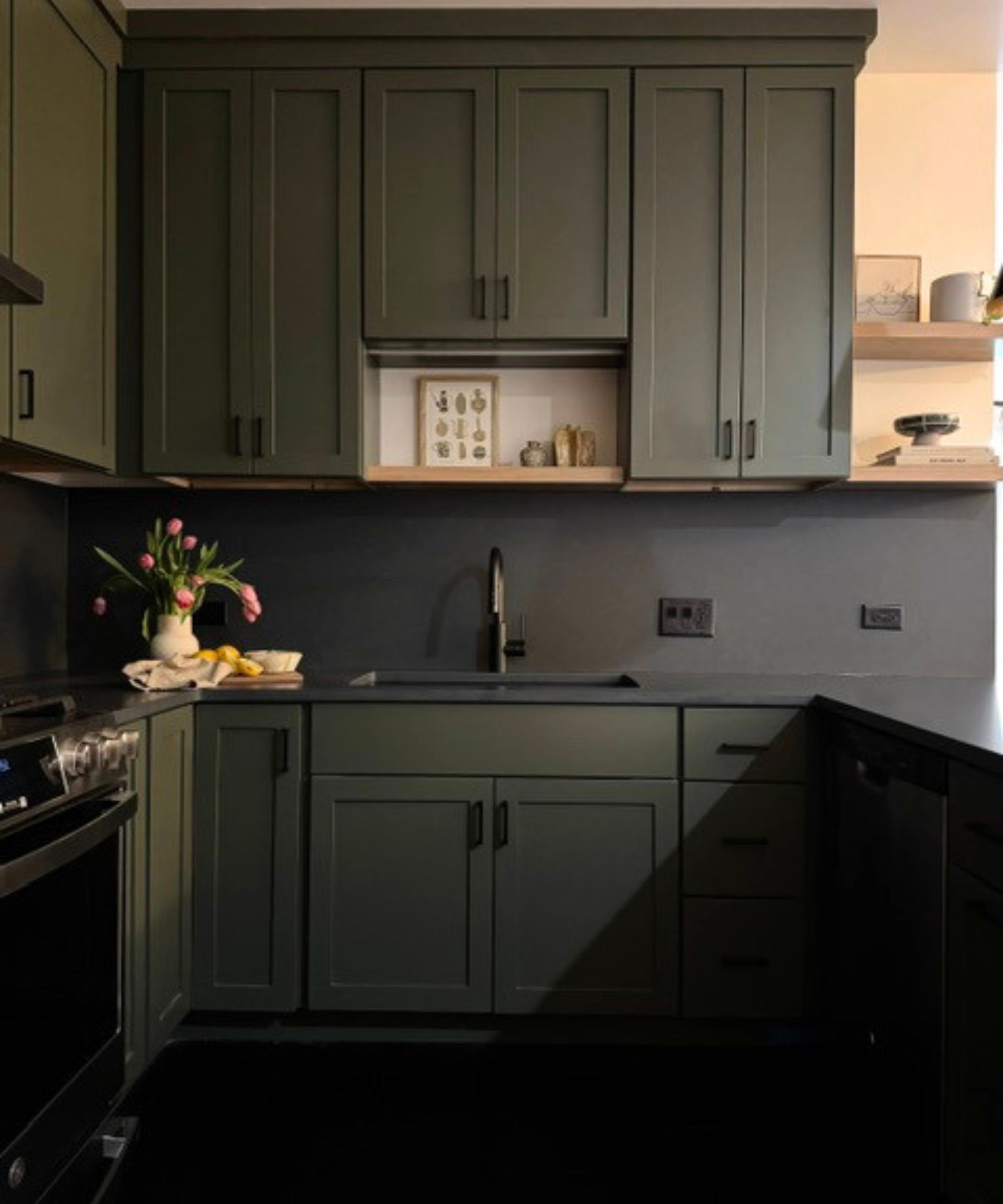 Cuisine avec armoires peintes Pewter Green de Sherwin-Williams, dosseret et comptoirs noirs, cuisinière, étagères ouvertes en bois avec céramiques et imprimés et vase de tulipes.