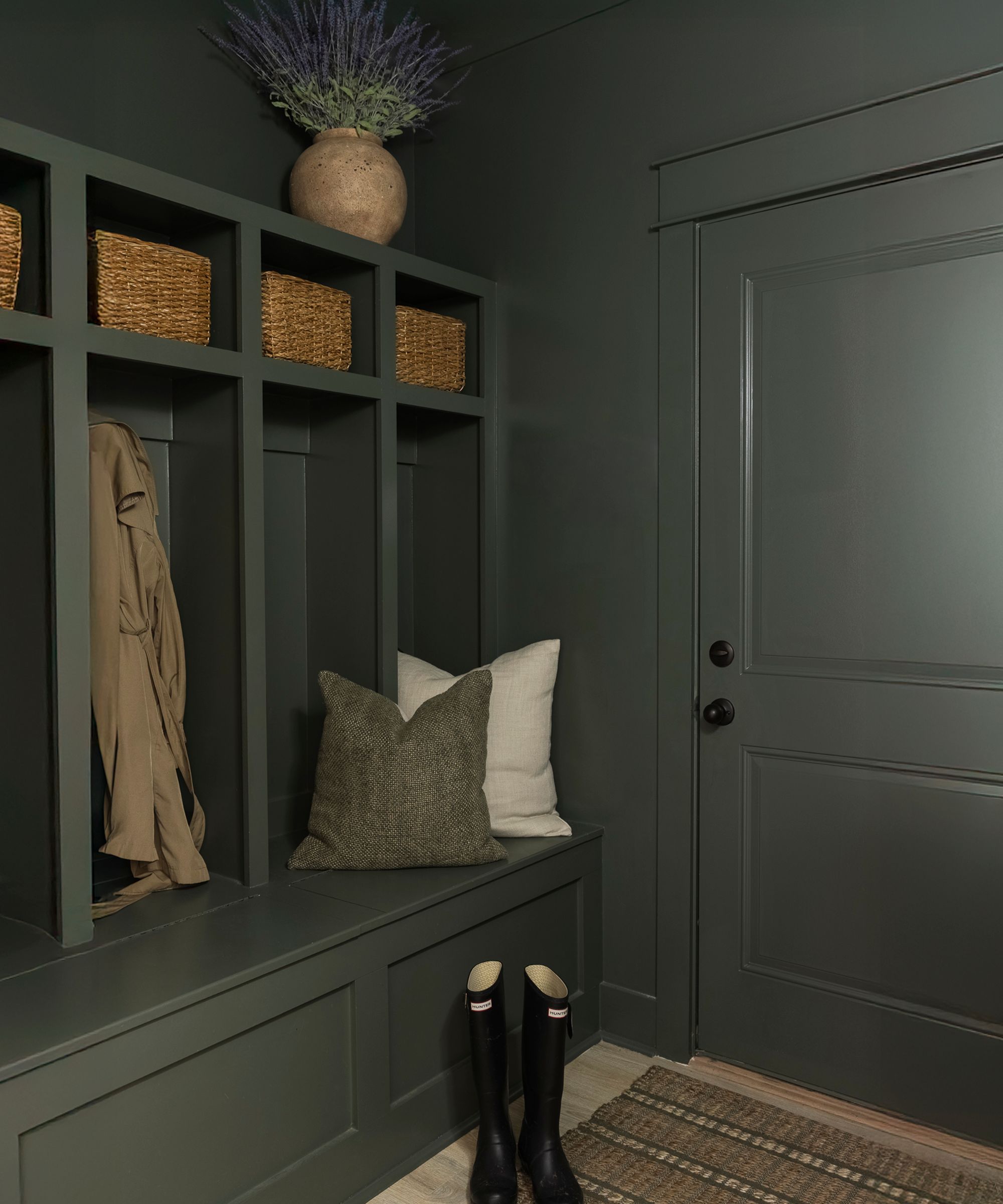 Vestiaire peint en vert Sherwin-Williams Pewter, avec un banc et des rangements intégrés, des paniers en rotin, un tapis en jute et une paire de Hunter Wellingtons.