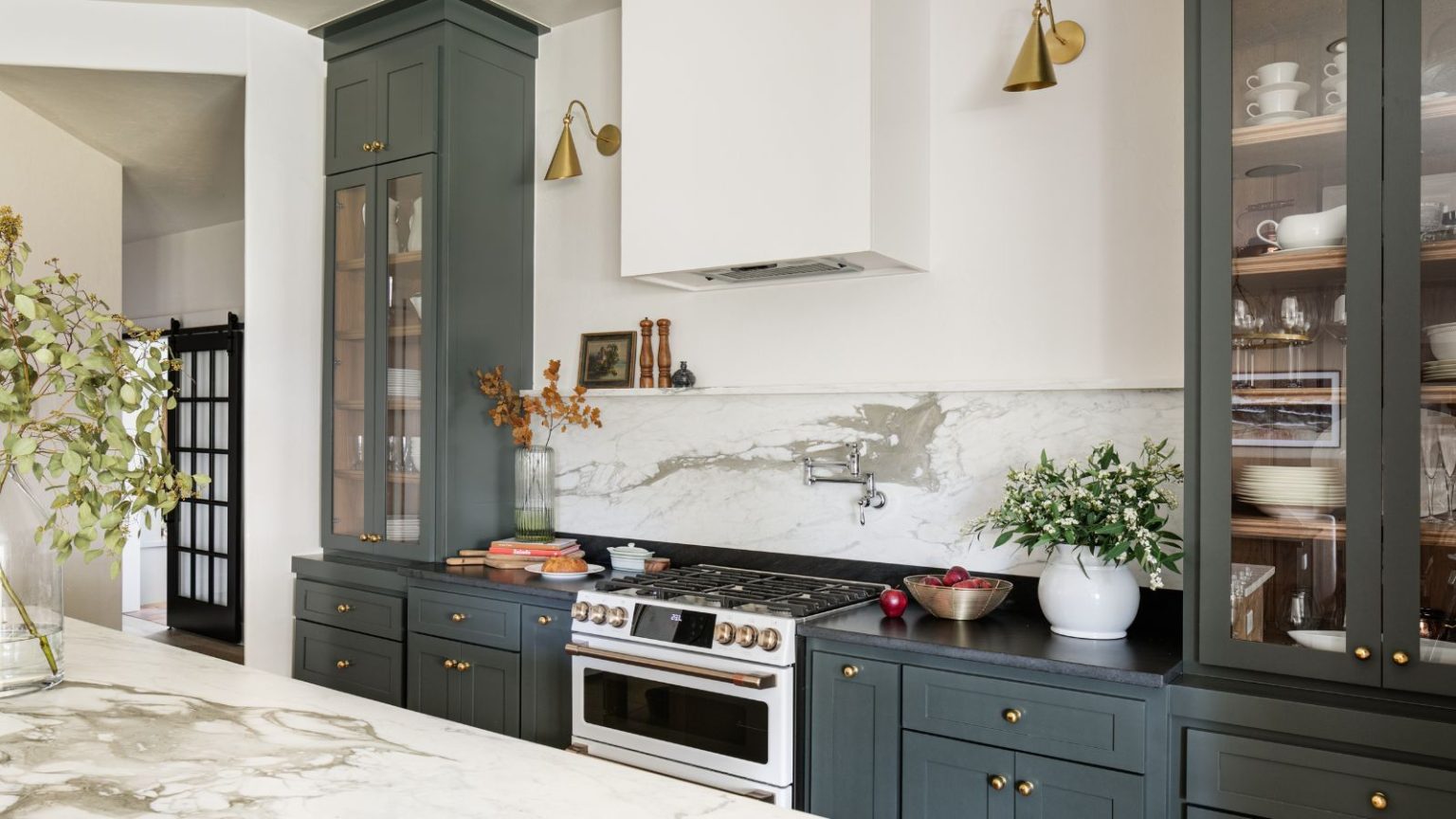 « Riche, invitant et un peu inattendu » – Pourquoi le vert étain de Sherwin-Williams reste la couleur de peinture préférée des designers Modern farmhouse style kitchen with marble backsplash, dark green cabinets painted in Sherwin-Williams Pewter Green, and glass-fronted cabinets filled with tableware.