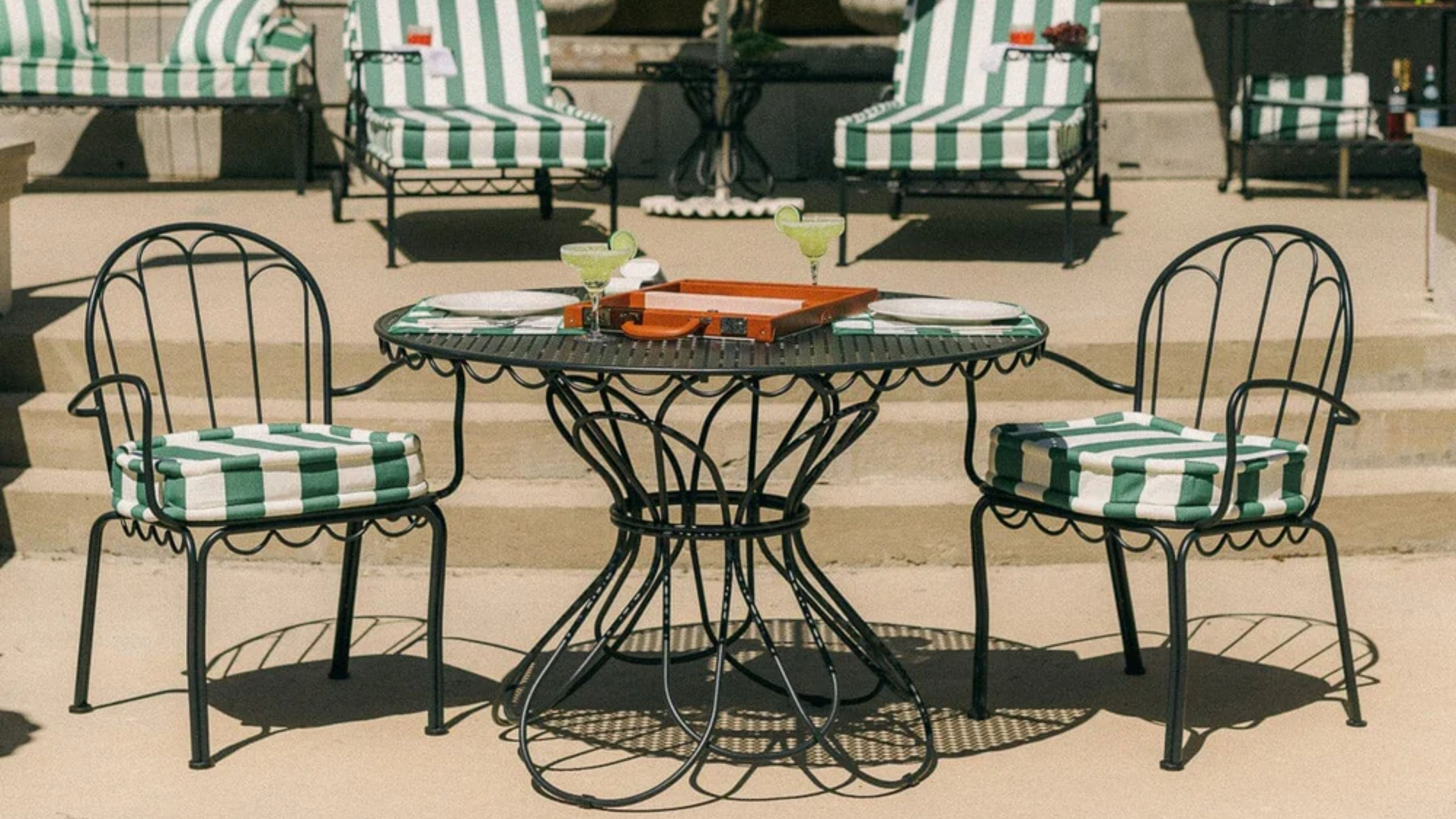 Table et chaises de bistro d'extérieur en fer fantaisistes, photographiées devant une rangée de chaises longues avec des coussins à rayures vertes et blanches
