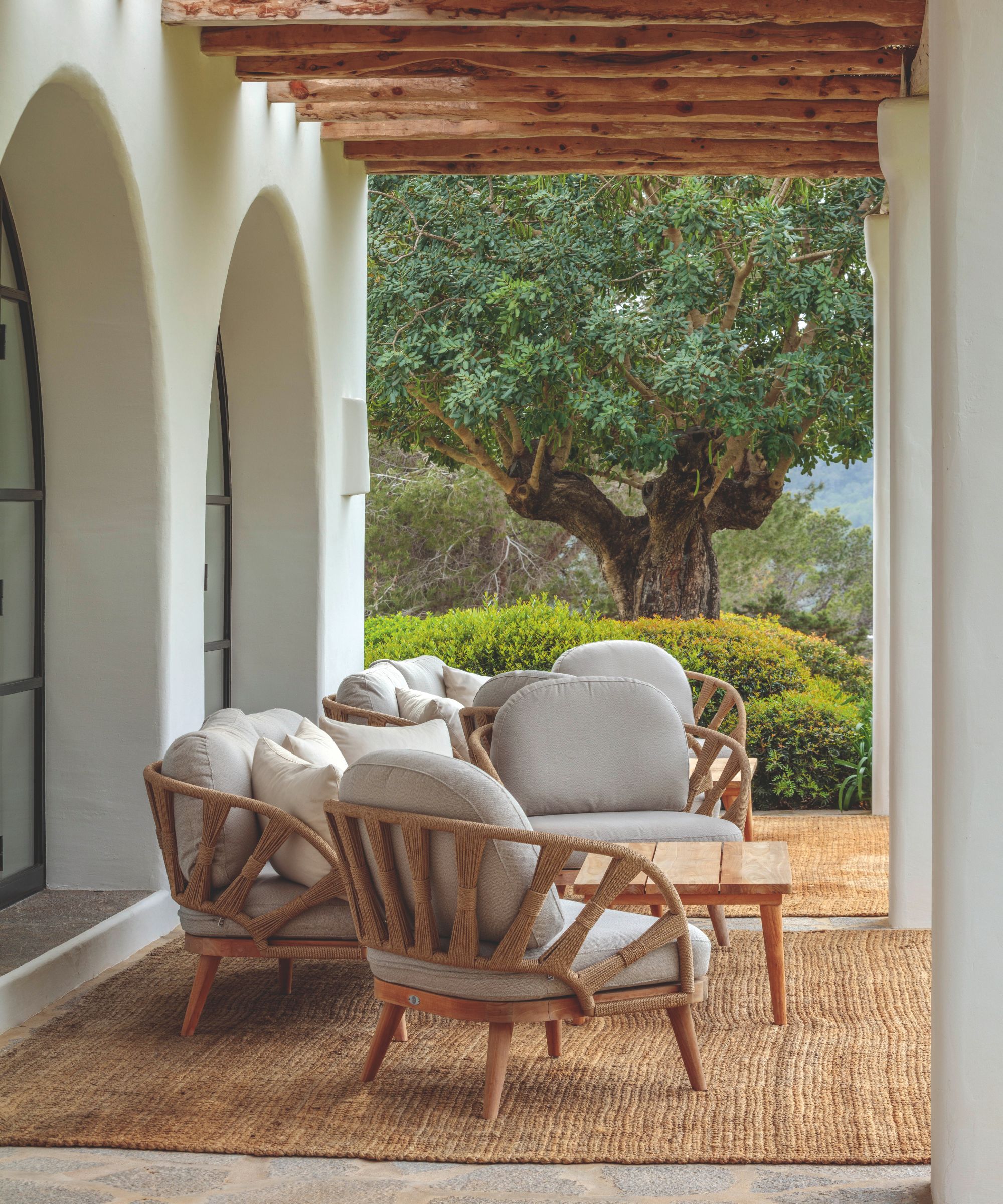 Patio avec poutres apparentes et murs voûtés blancs, tapis en jute au sol, chaises longues en rotin et table en bois, donnant sur un jardin méditerranéen.