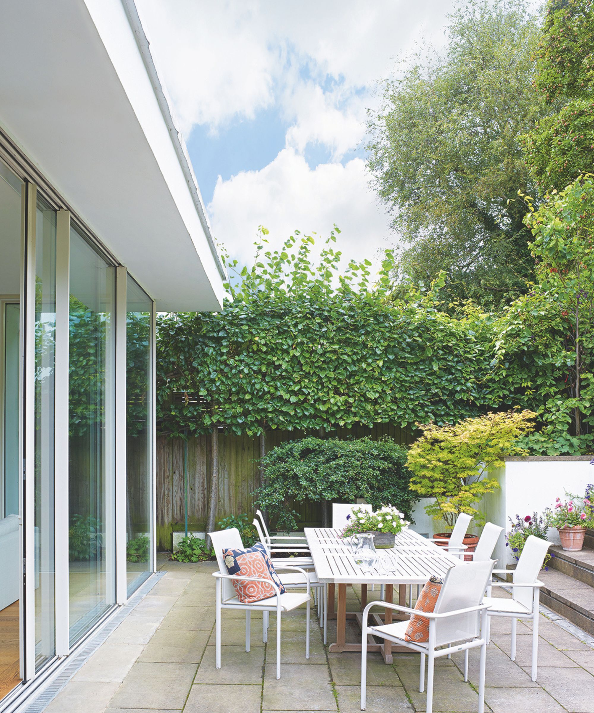 Maison moderne avec portes vitrées donnant sur un patio avec un ensemble de salle à manger en métal blanc comprenant une longue table et des chaises, et des plantations d'intimité avec des haies et des arbres.